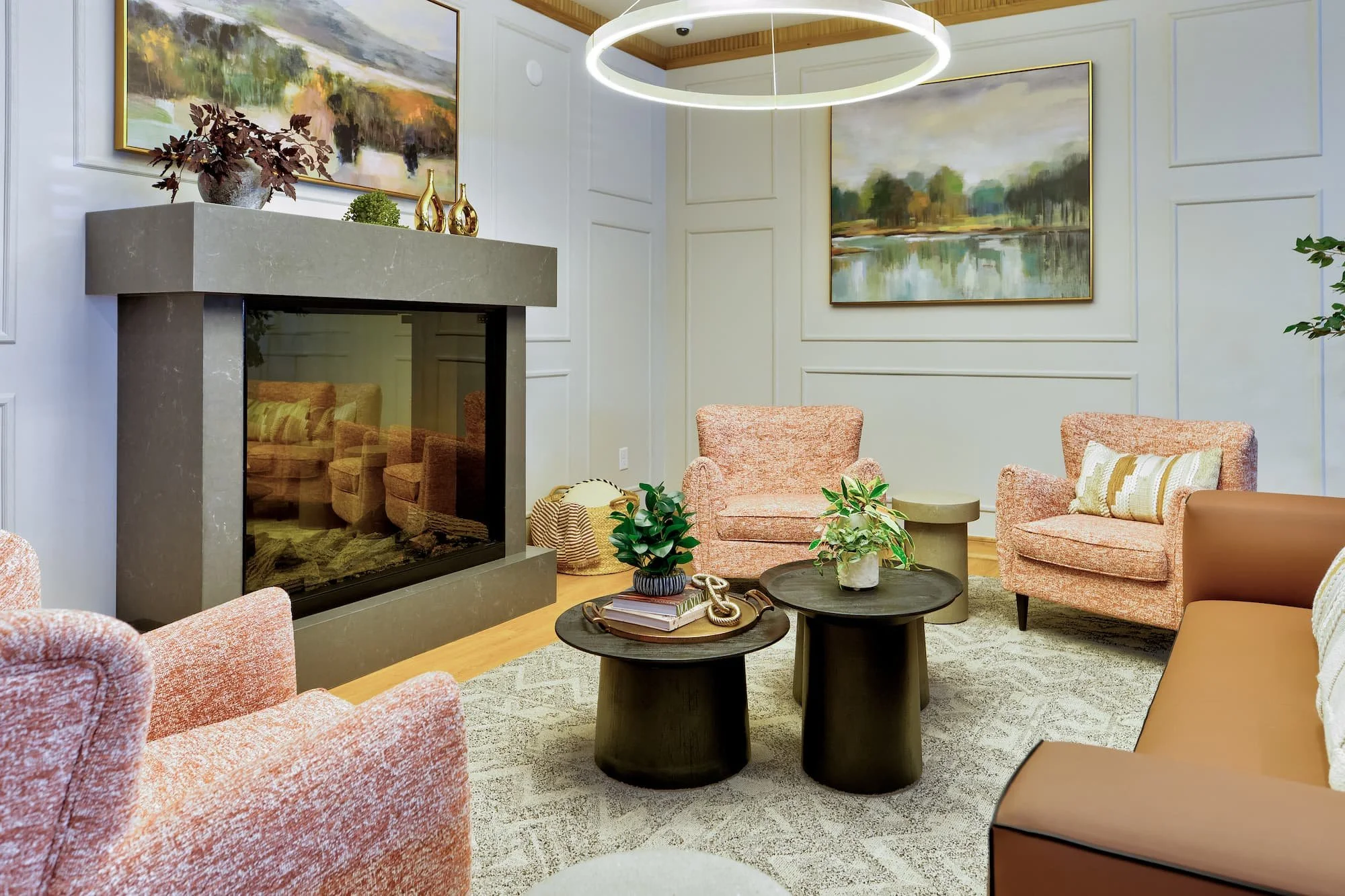 Cozy living room with a modern fireplace, upholstered pink chairs, and framed landscape paintings on paneled walls. Round coffee tables with decor, and a circular light fixture hanging from the ceiling. Amenity space at Birch & Branch in Raleigh.