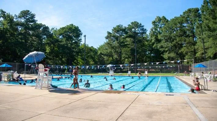 Public outdoor swimming pool with people swimming and relaxing under umbrellas on a sunny day, surrounded by trees in Raleigh, NC