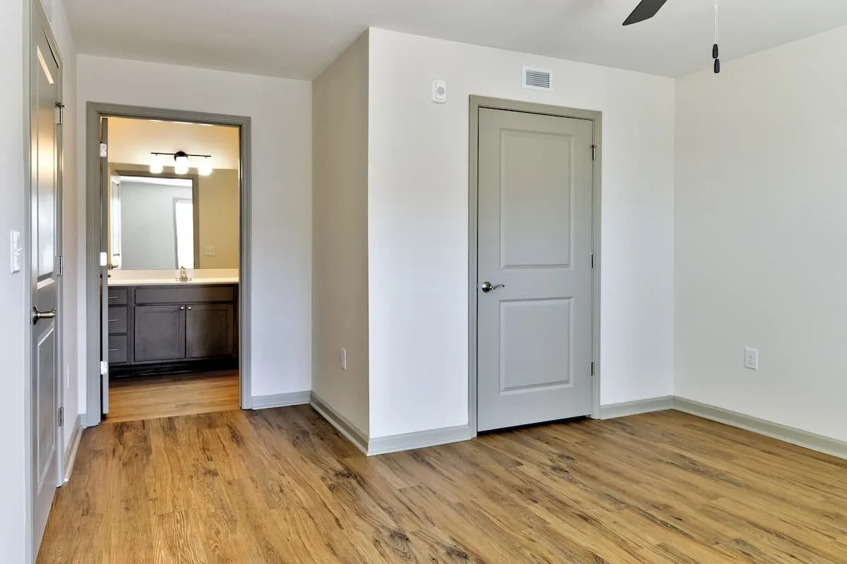 Empty room with wood flooring, white walls, two doors, and a ceiling fan, with a view into a bathroom with dark cabinets and a mirror at Birch & Branch Apartments in Raleigh, NC.