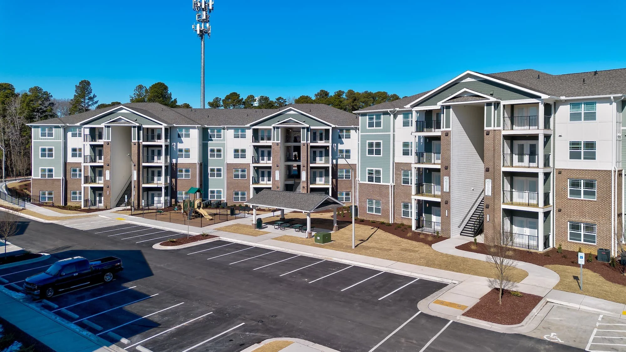 A modern multi-story apartment complex with a parking lot in the foreground, a playground and picnic area, and surrounded by trees under a clear blue sky. Birch & Branch Apartments in Raleigh, North Carolina