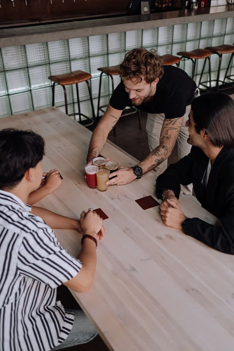 A bartender serving drinks to two customers seated at a bar counter in a modern bar or cafe.