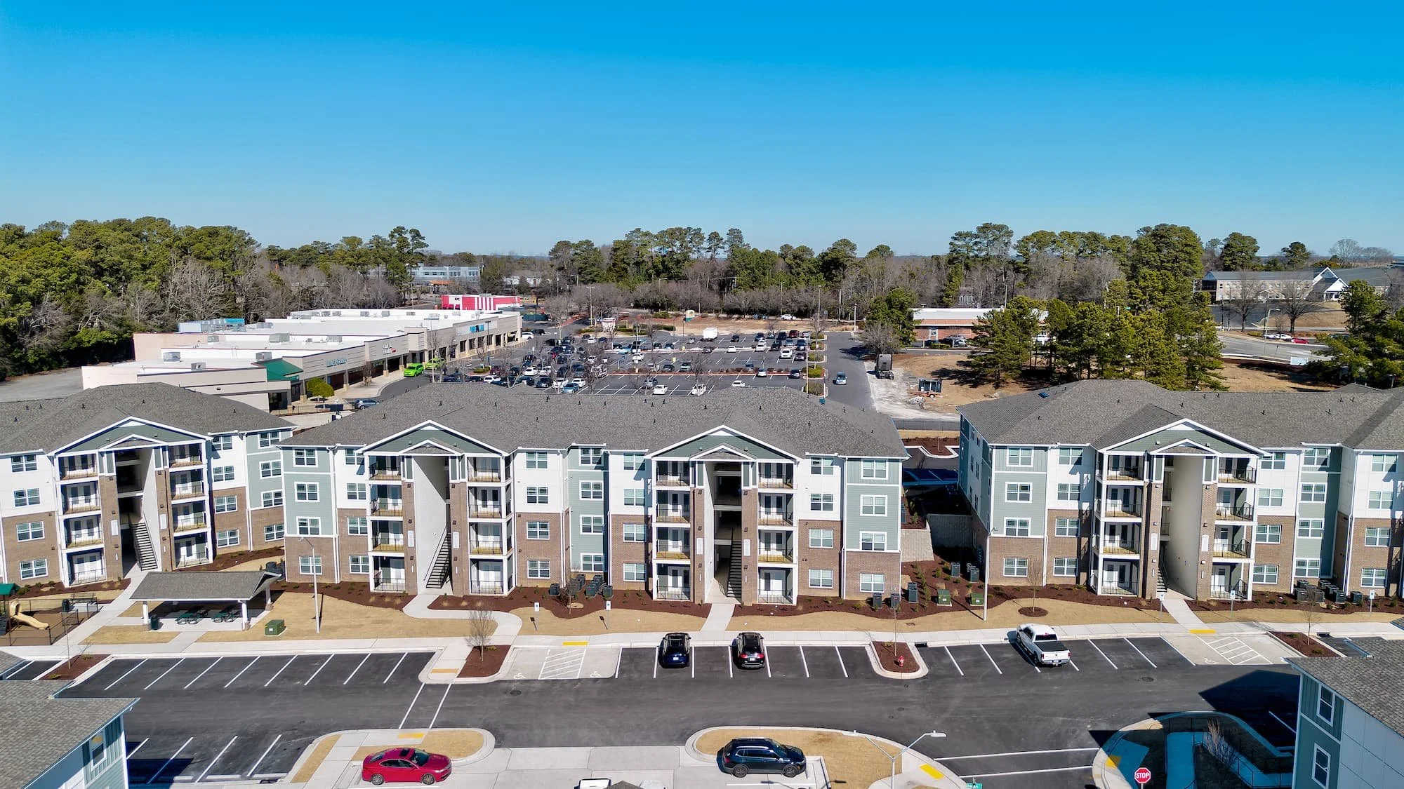 An aerial view of Birch & Branch apartment complex in Raleigh, NC with parking spaces, neighboring commercial buildings, and a wooded area with trees under a clear blue sky.