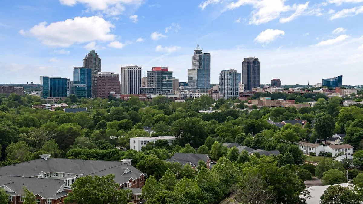 Skyline of the city of Raleigh, NC with skyscrapers, surrounded by lush green trees and residential houses in the foreground.