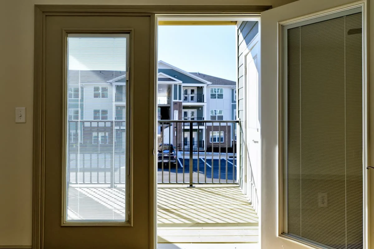 Interior view of a door opening at Birch & Branch Apartments to a small outdoor balcony with a railing, overlooking a parking lot and neighboring apartment buildings under a clear blue sky.