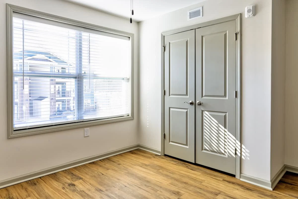 Empty room with a large window with blinds, hardwood floor, and a closed closet door. Sunlight creates a shadow pattern on the closet door at Birch & Branch Apartments in Raleigh, NC.