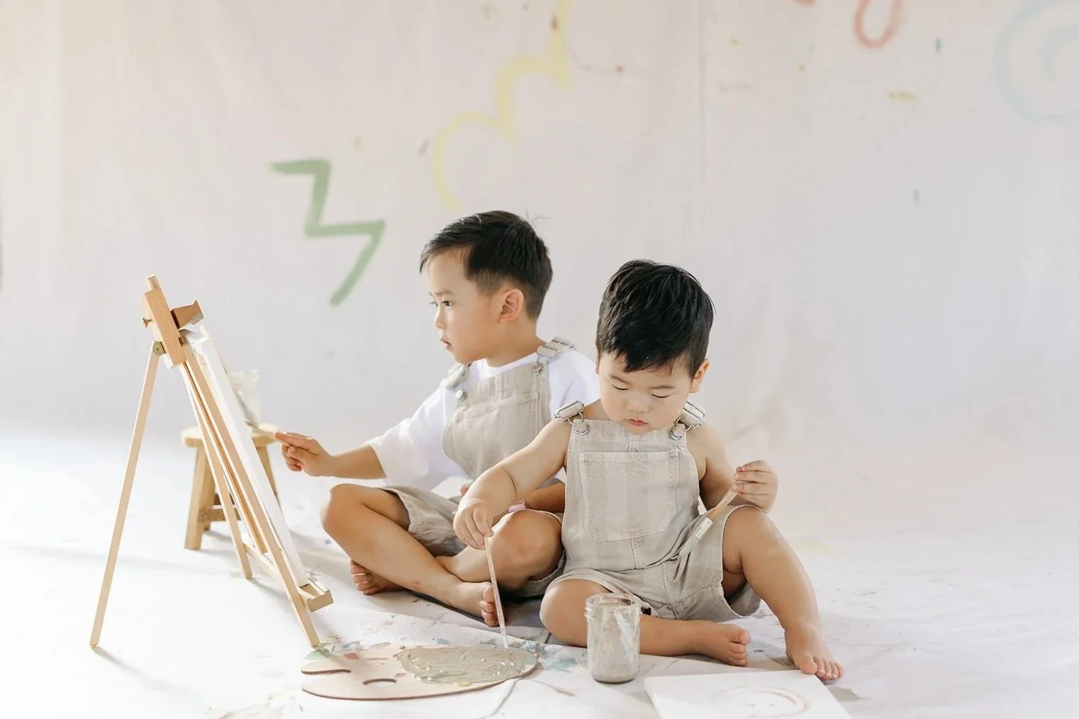 Two young children, a boy and a toddler, sit on a white floor painting with brushes and watercolors on paper, with an easel and paint supplies nearby.