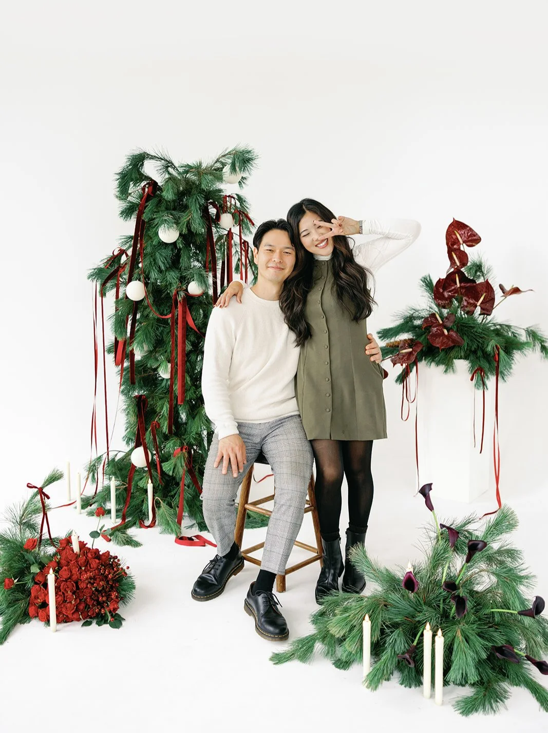 Two people smiling and posing with Christmas decorations. The man is sitting on a stool, and the woman is standing next to him making a peace sign. There are decorated Christmas trees, flowers, candles, and red ribbons in the background.