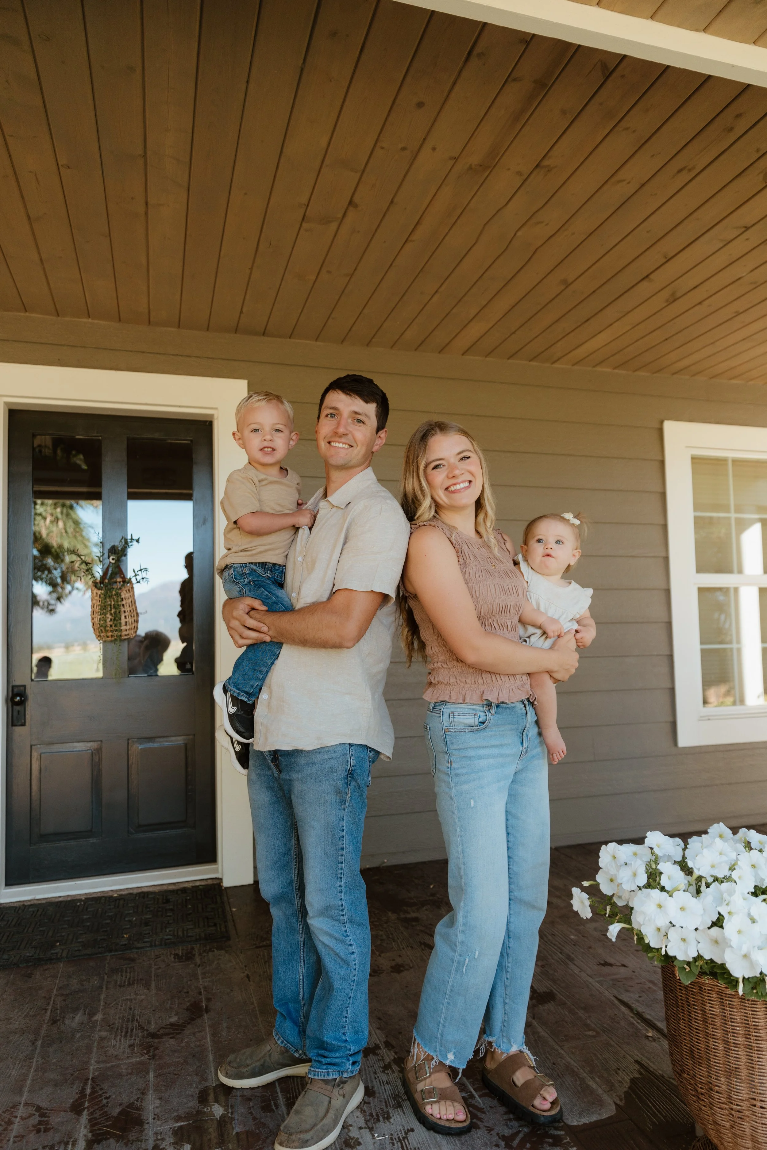 A family of four standing outside their house, smiling at the camera. The father is holding a young boy, and the mother is holding a young girl. The house has a wooden ceiling and a door with glass panels, and there is a basket of white flowers in the foreground.