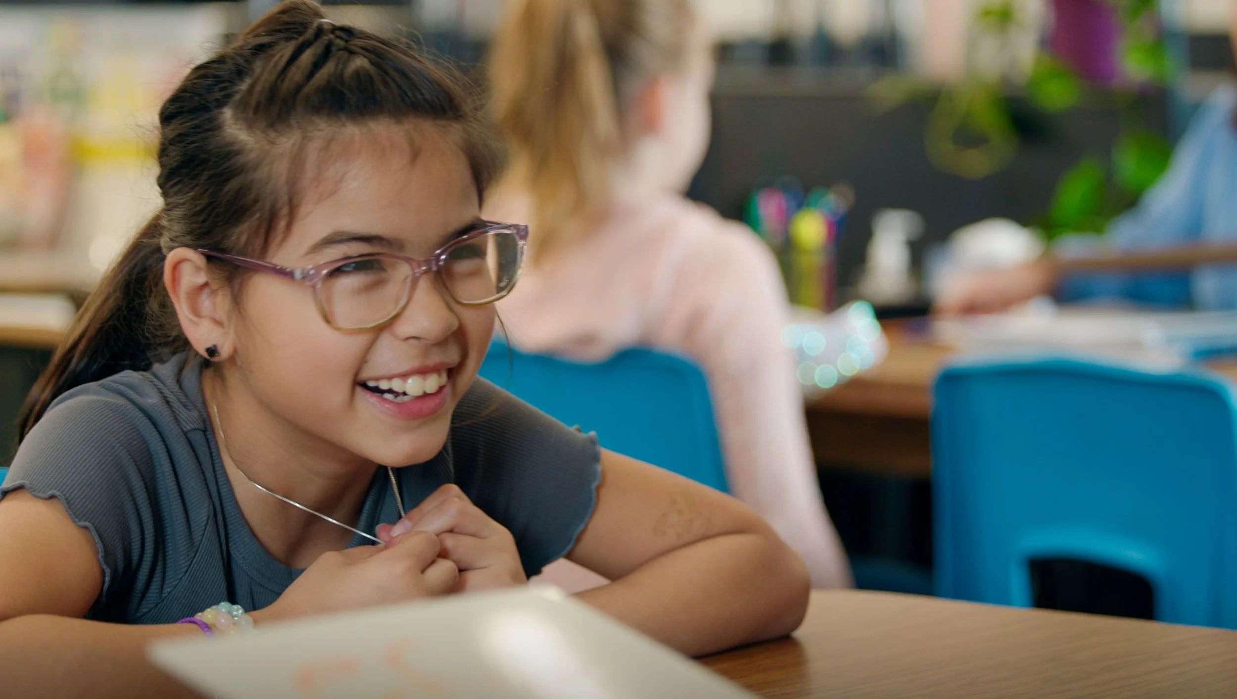 Smiling student wearing glasses sits at a classroom desk with classmates in the background
