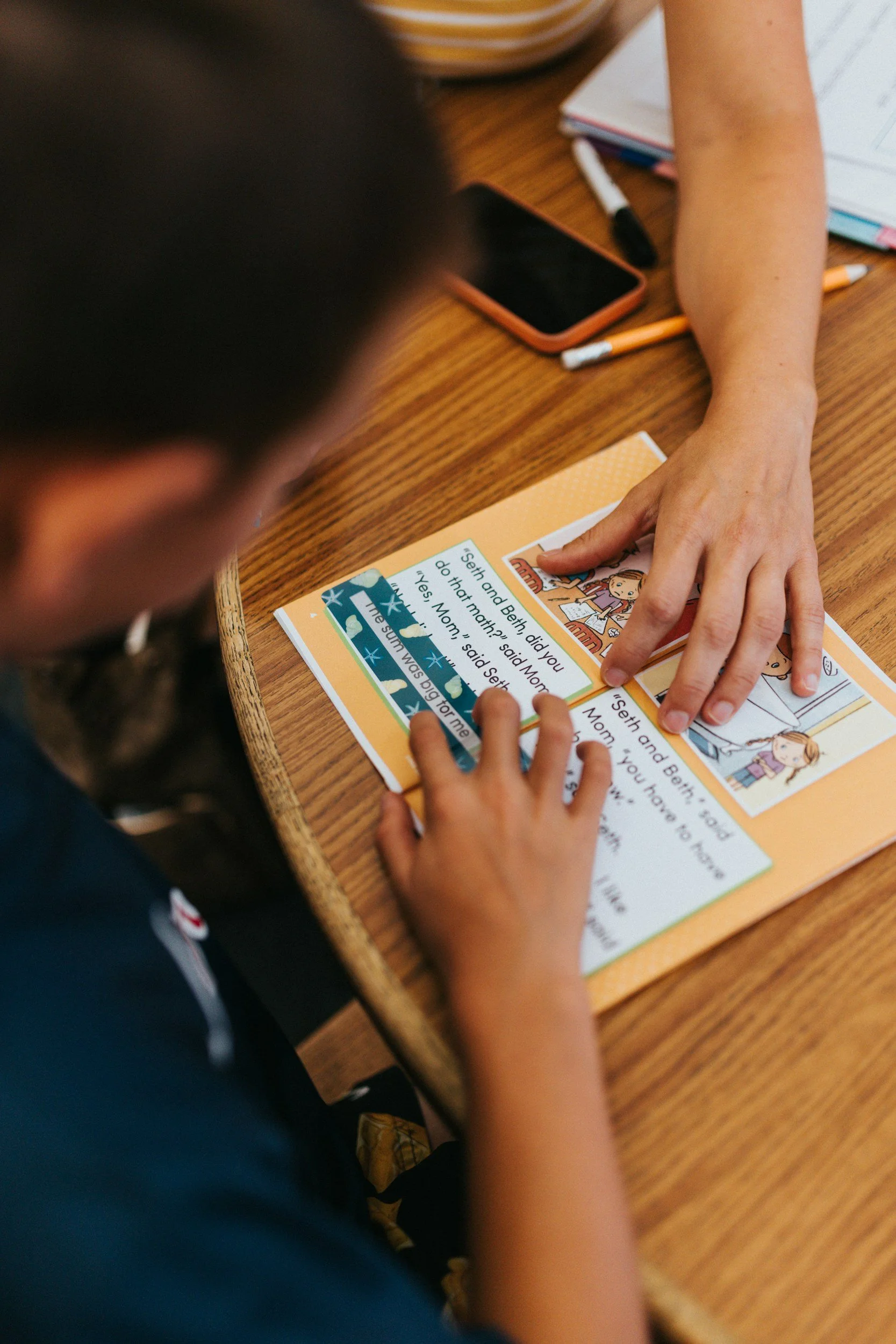 Adult helping a child read an illustrated workbook at a wooden table with a smartphone and pencils nearby
