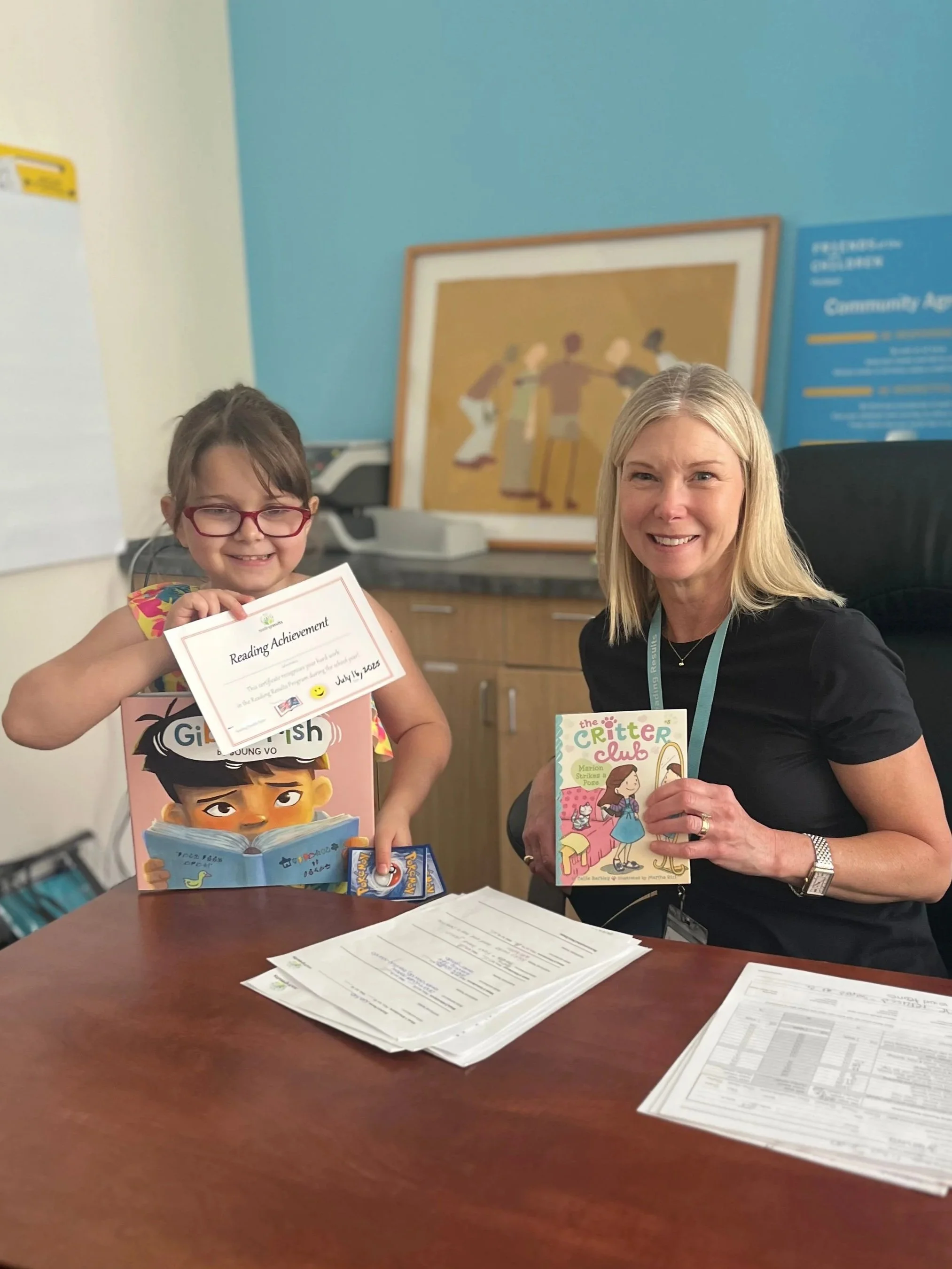 Child holding a reading achievement certificate and book beside a teacher holding a children's book in a classroom