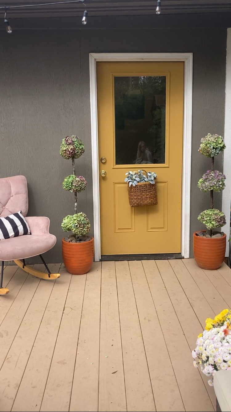 Decorative porch area with yellow door, potted plants, pink rocking chair with striped pillow, and a bunch of flowers in a white container.