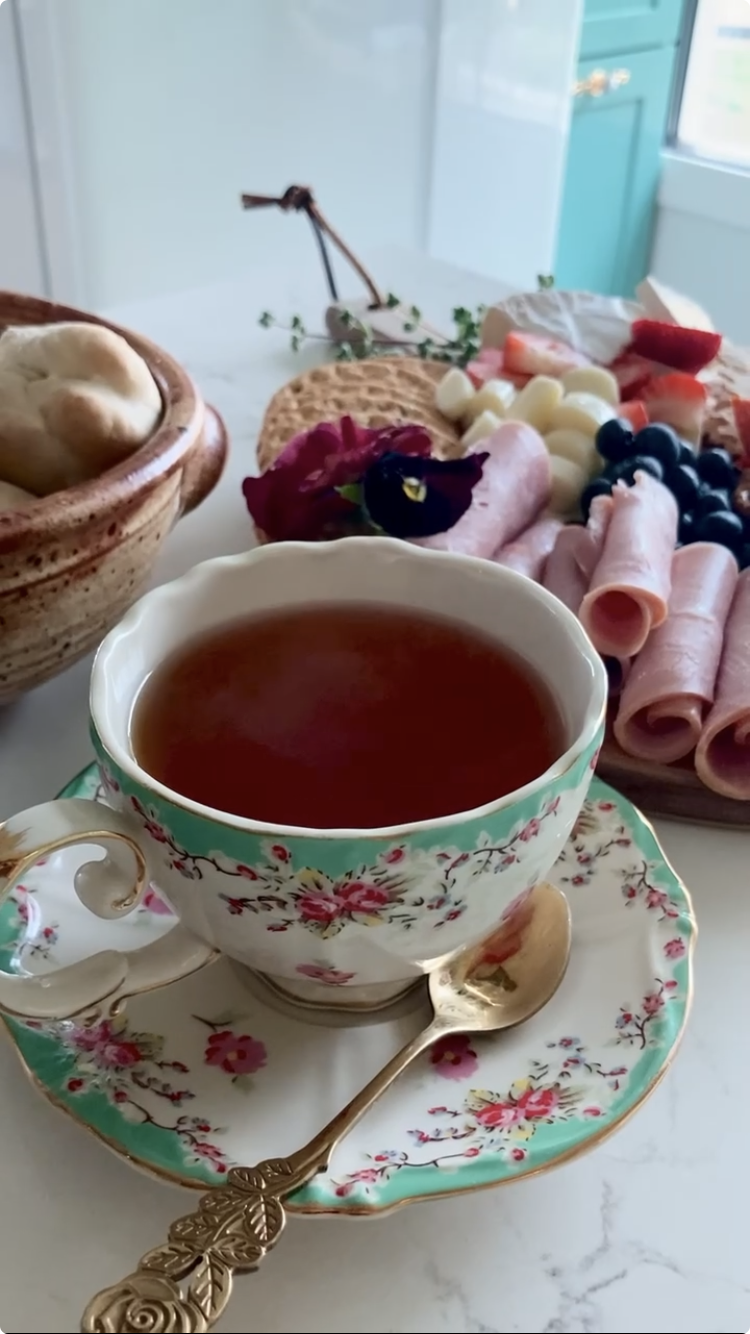 A cup of tea on a matching floral saucer with a gold spoon, set on a white marble surface amidst an assortment of breakfast foods including bread, sliced meats, cheese, berries, and sliced watermelon, with a rustic and cozy kitchen background.