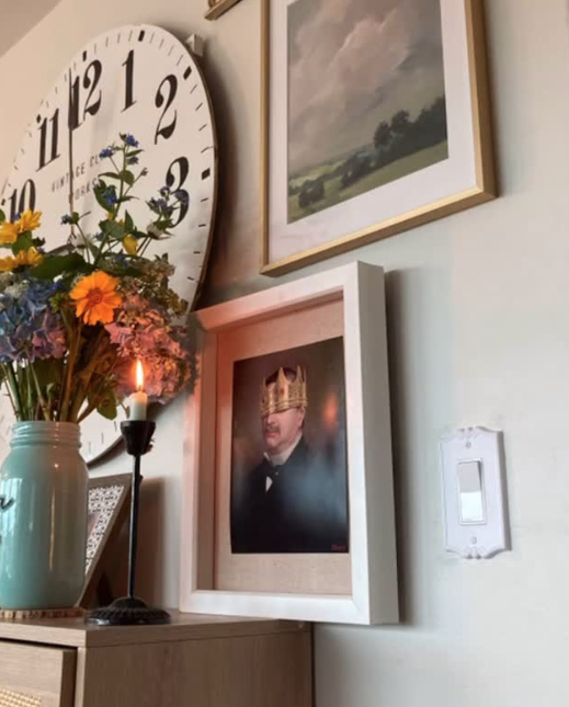 A corner of a room with a large wall clock, a framed landscape painting, a bouquet of colorful flowers in a vase, a lit candle in a holder, and a framed photograph of a man wearing a crown and glasses, all on top of a wooden surface.