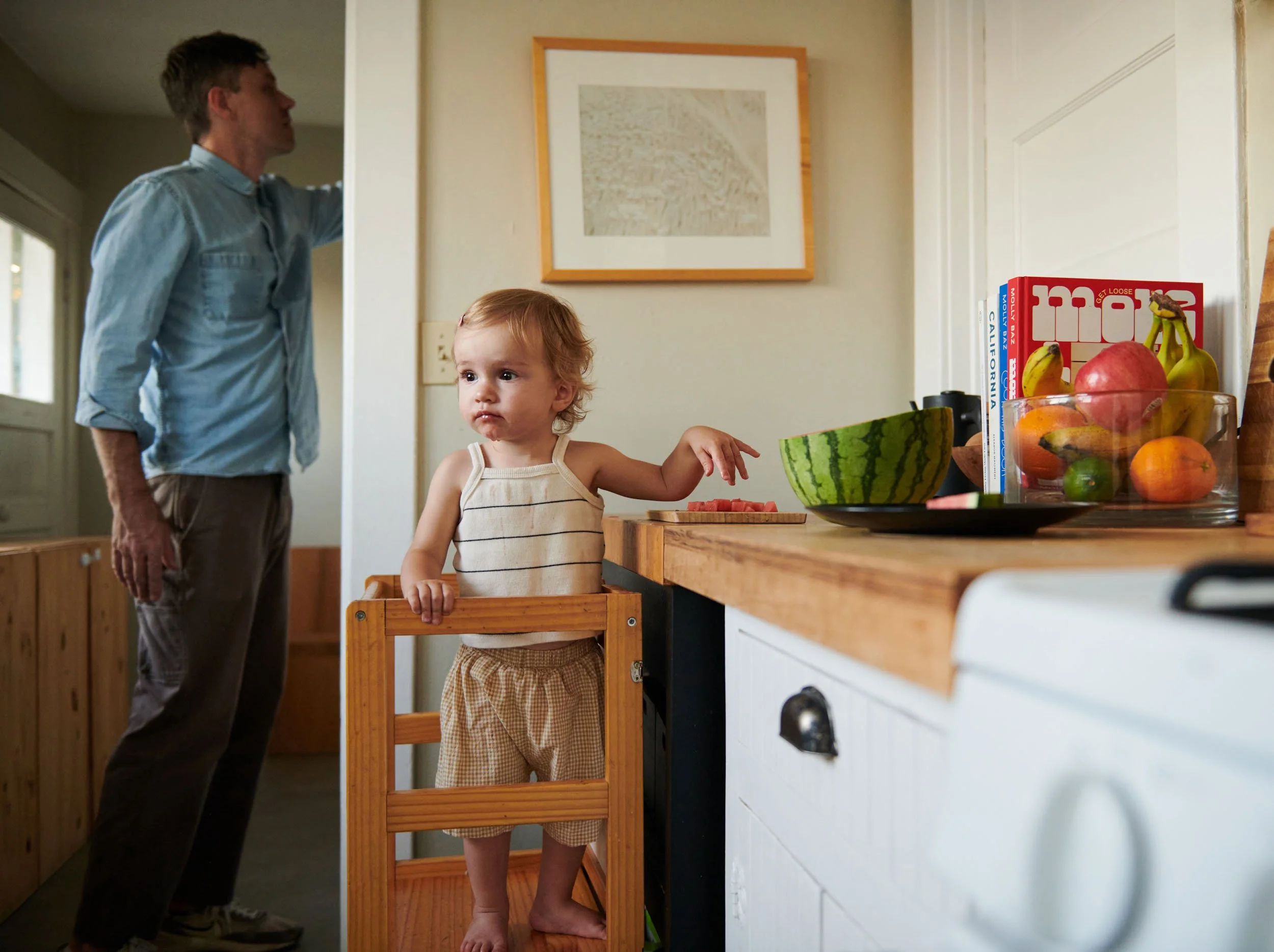 Toddler standing at a learning tower by the kitchen counter while dad reaches for a cupboard, documentary family photography in Los Angeles