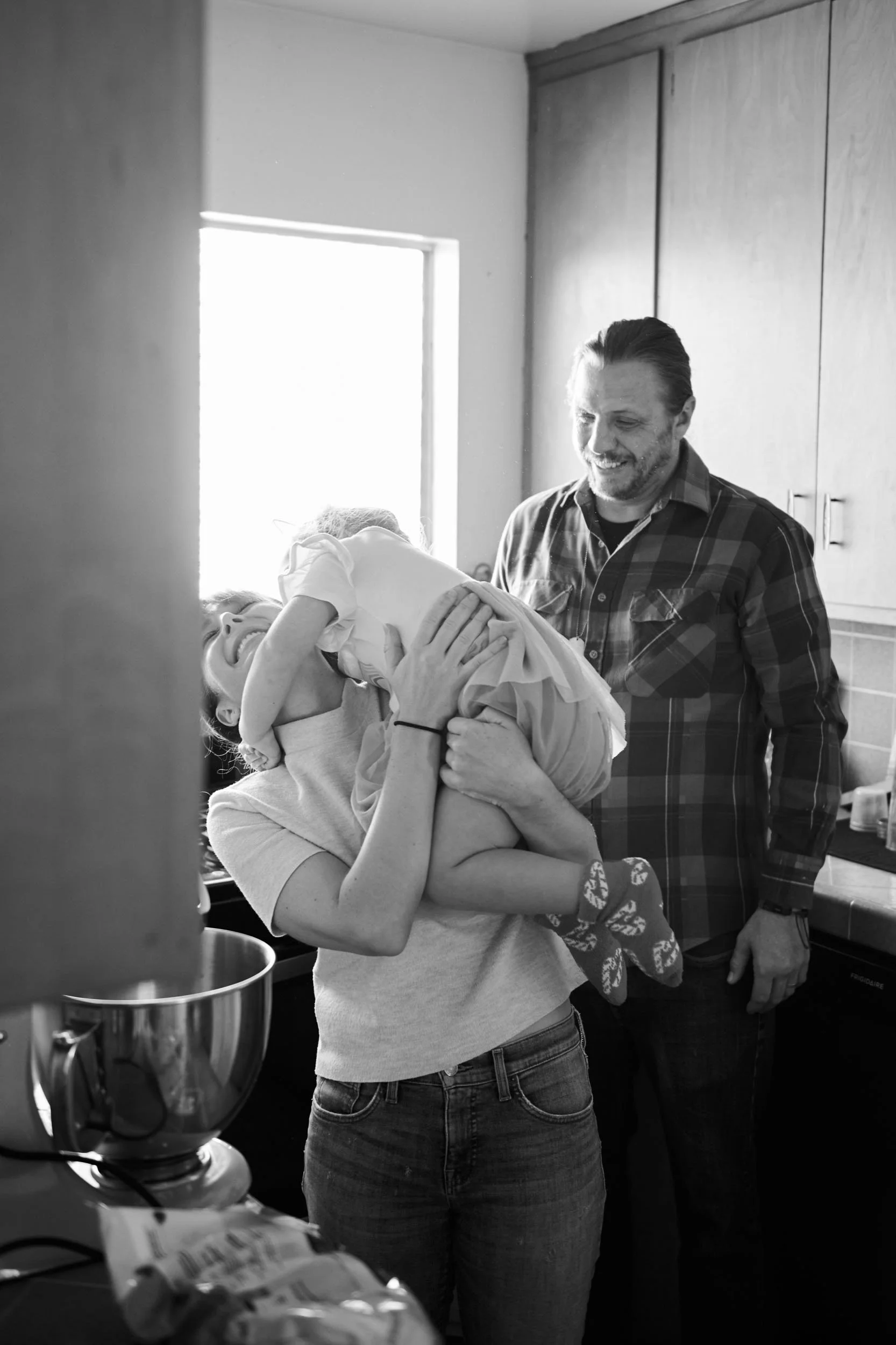 Black and white photo of a mom lifting her laughing daughter while dad smiles nearby at home, documentary family photography Los Angeles