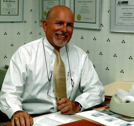 A smiling man with a bald head, beard, and glasses on a chain, dressed in a white shirt and yellow tie, sitting at a desk in an office with framed certificates on the wall.