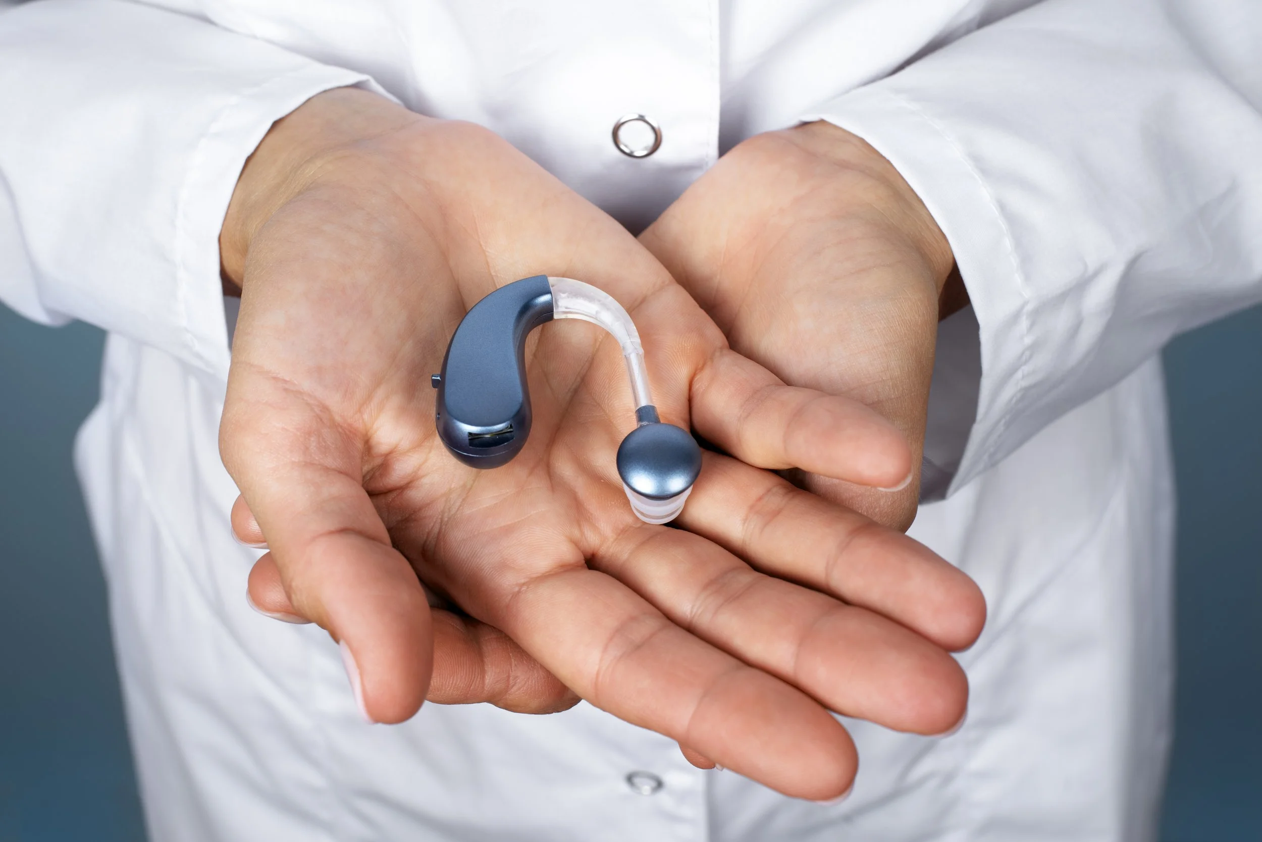 A healthcare professional holding a hearing aid in their hand, with part of the device visible on the palm, wearing a white lab coat.