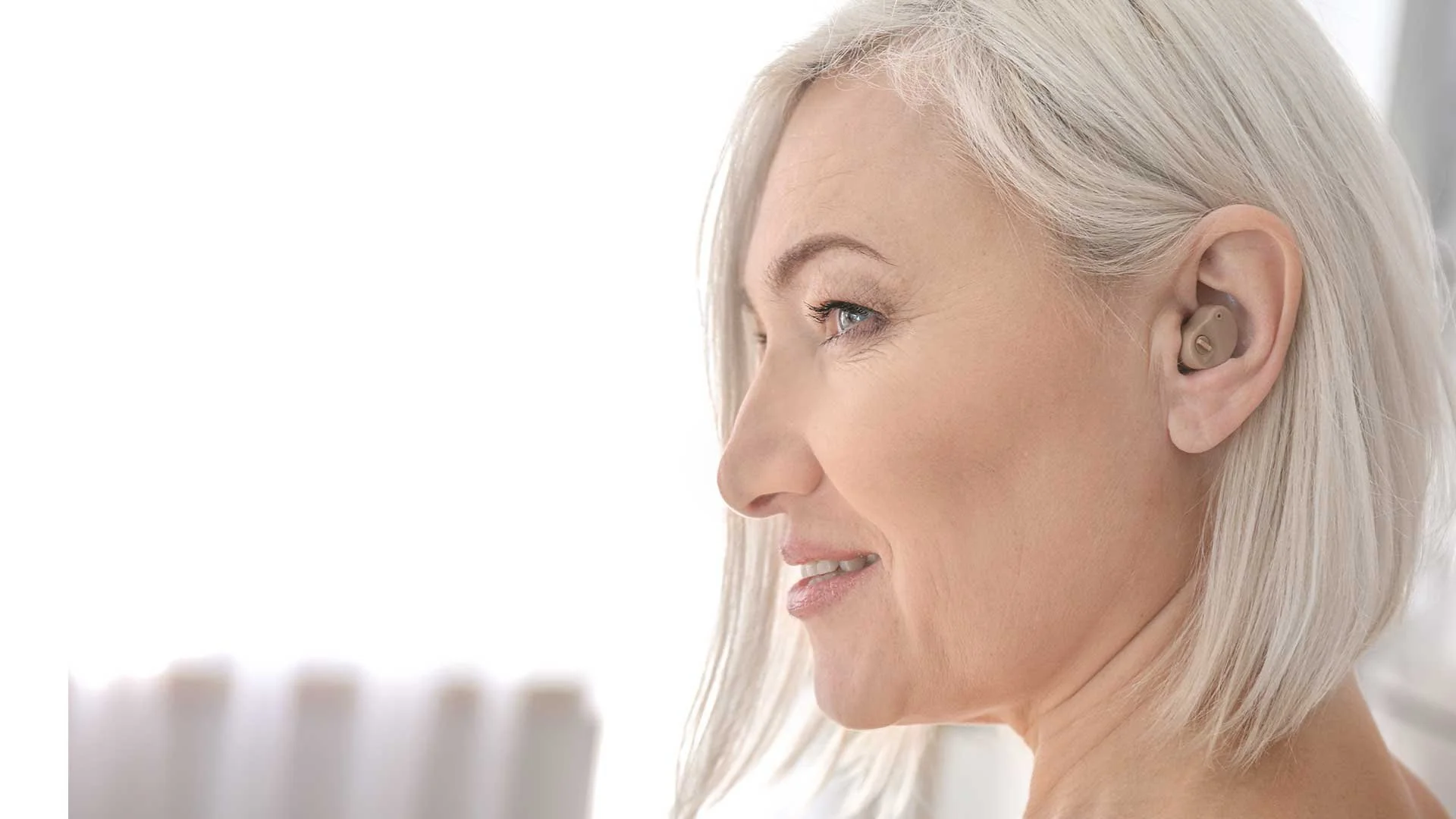 Close-up of a smiling middle-aged woman with long blonde hair wearing a beige hearing aid in her right ear.