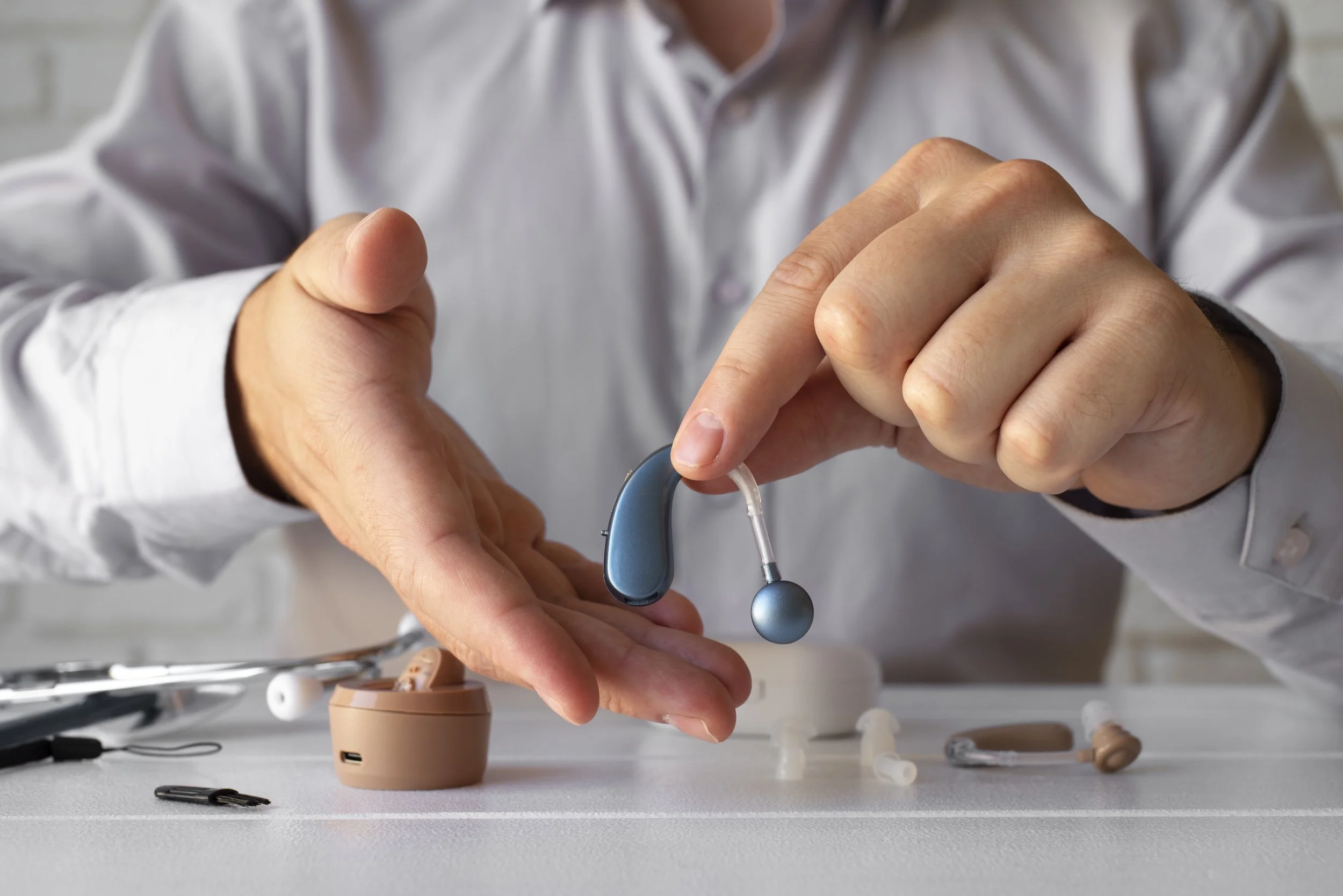 A person in a white shirt is handling a blue behind-the-ear hearing aid, with various medical devices and accessories on a white surface in front of them.