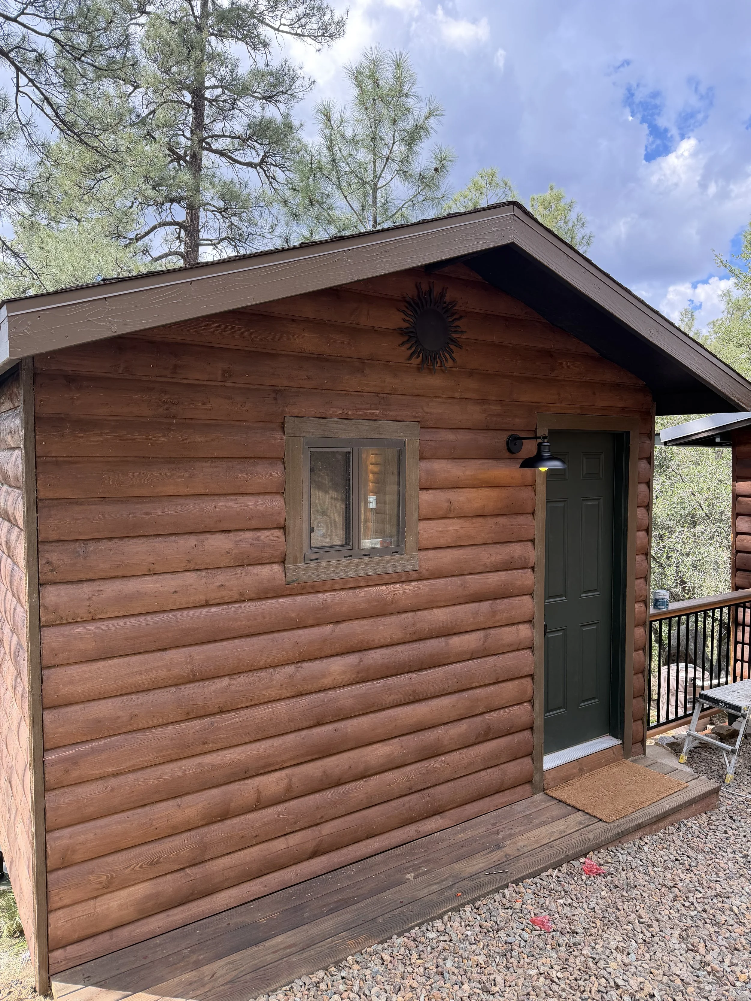 A small wooden cabin with a green door, a window, and a black outdoor light fixture.