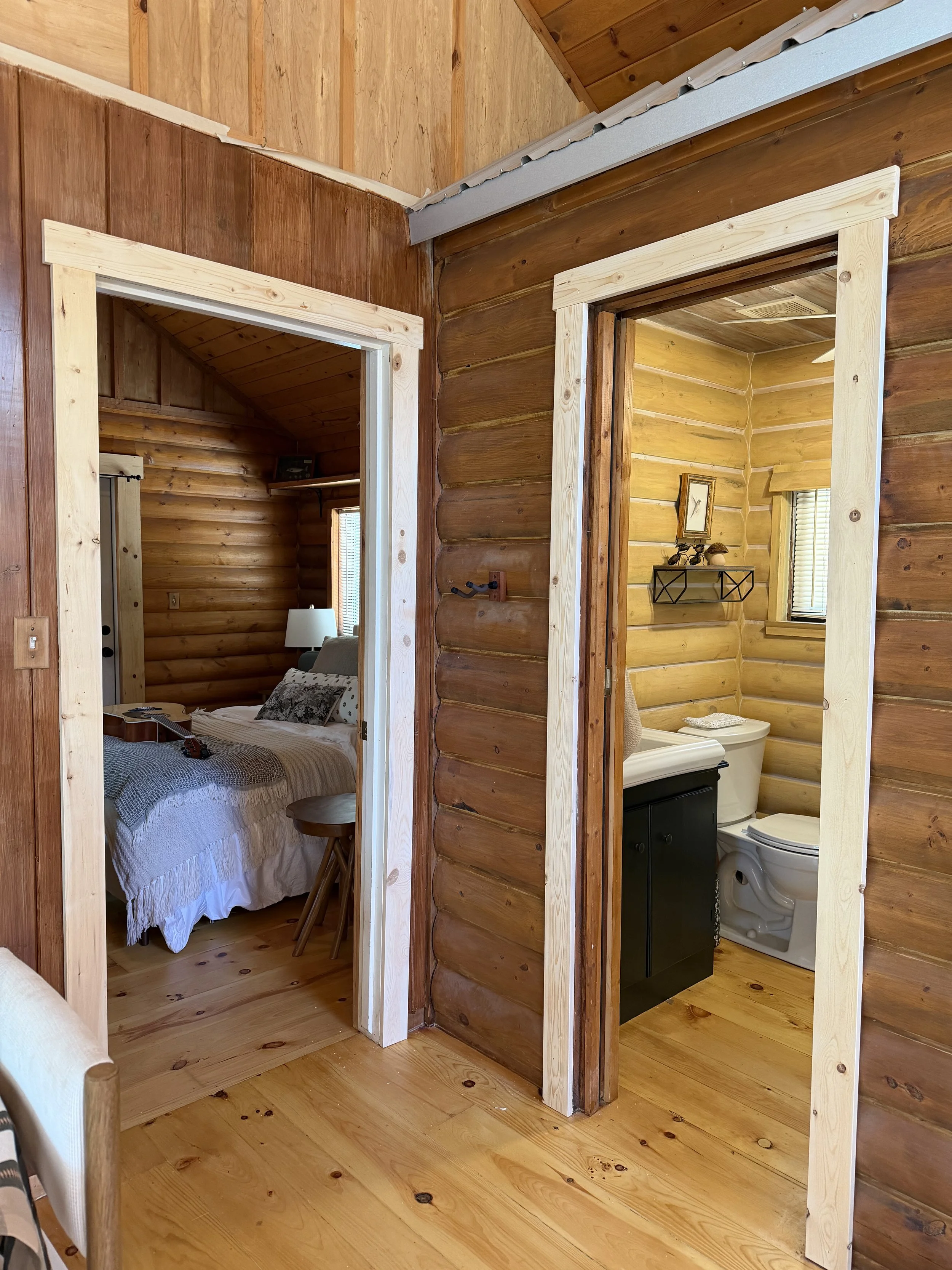 Interior of a wooden cabin with two doorways: one leads to a bedroom with a bed, pillows, nightstand, and lamp; the other leads to a bathroom with a toilet, black vanity, and framed picture on the wall.