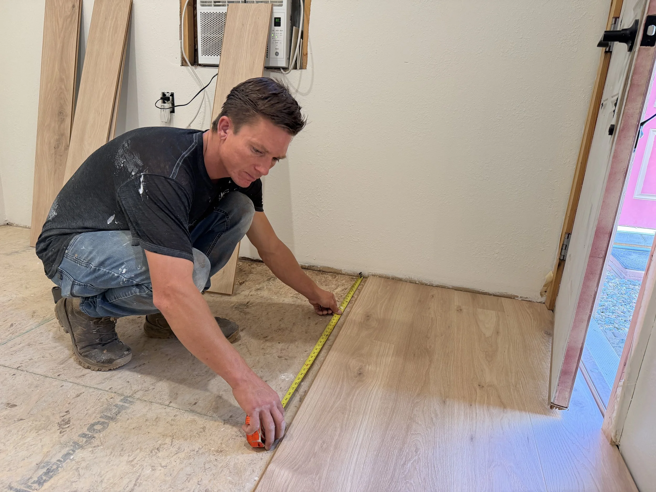 A man crouches down measuring wooden flooring with a tape measure inside a home renovation site. Wooden planks and construction materials are visible around him.
