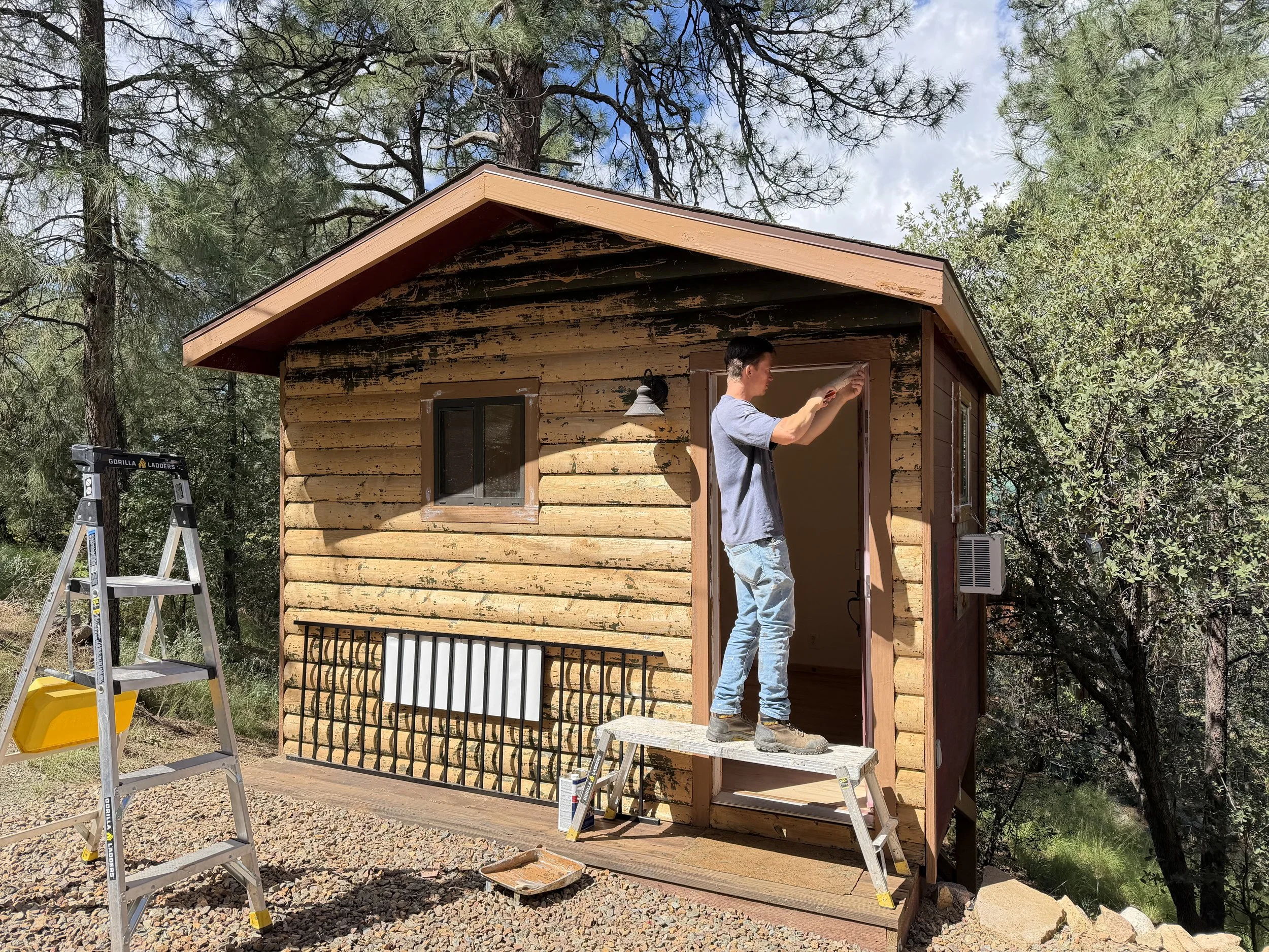 A man is standing on a step stool working on the exterior of a small wooden house, with trees and a cloudy sky in the background. He appears to be applying paint or stain to the house's trim.
