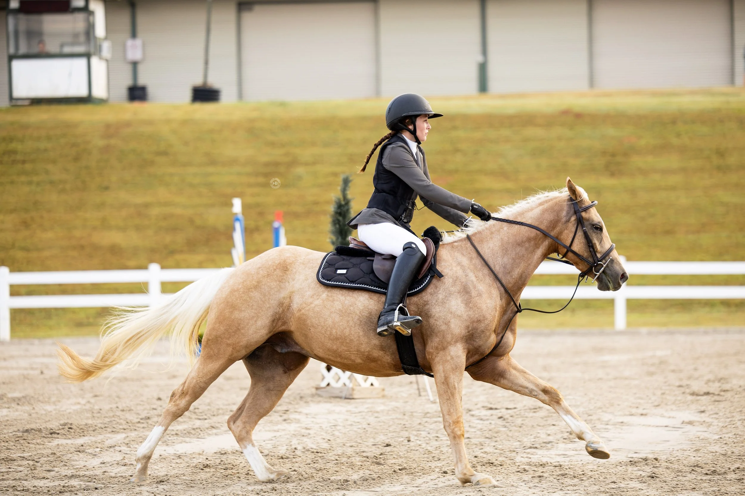 A female rider in equestrian attire riding a palomino horse during a jumping competition, with a blurred background of equestrian facilities.