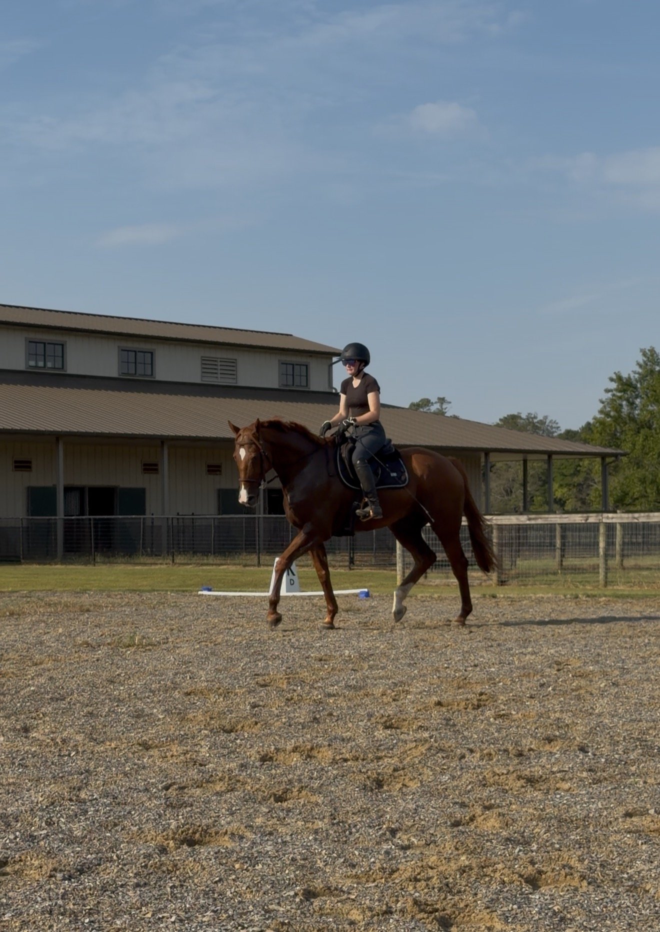 A woman wearing a black helmet and dark clothing is riding a brown horse with a white blaze on its face. They are in an outdoor riding arena with a building in the background.