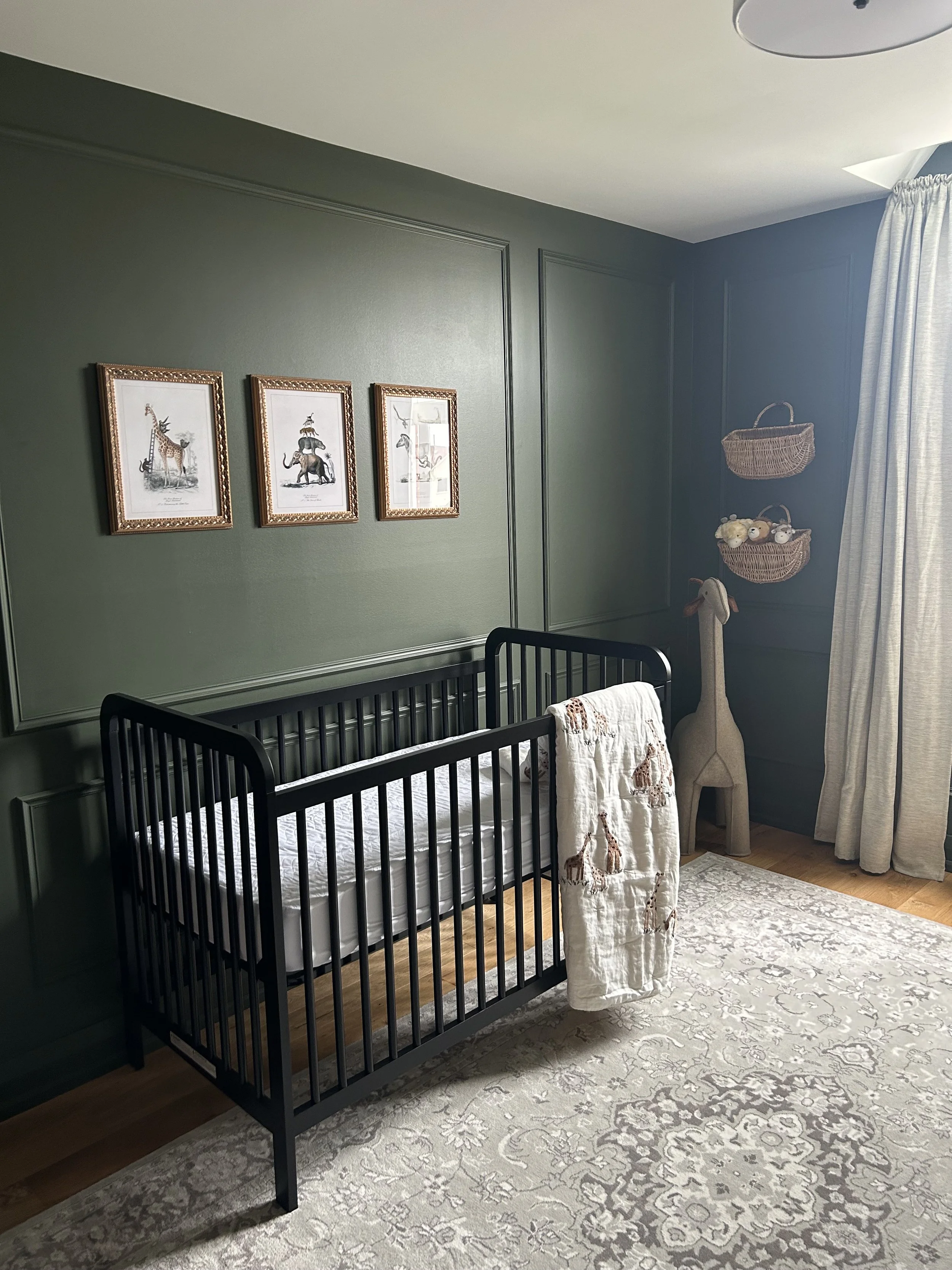 A nursery with a black crib, wall art of animals, wicker baskets on the wall, a giraffe-shaped stuffed animal, and a cream-colored quilt with giraffes on it. There's a patterned rug on the wooden floor and white curtains by the window.