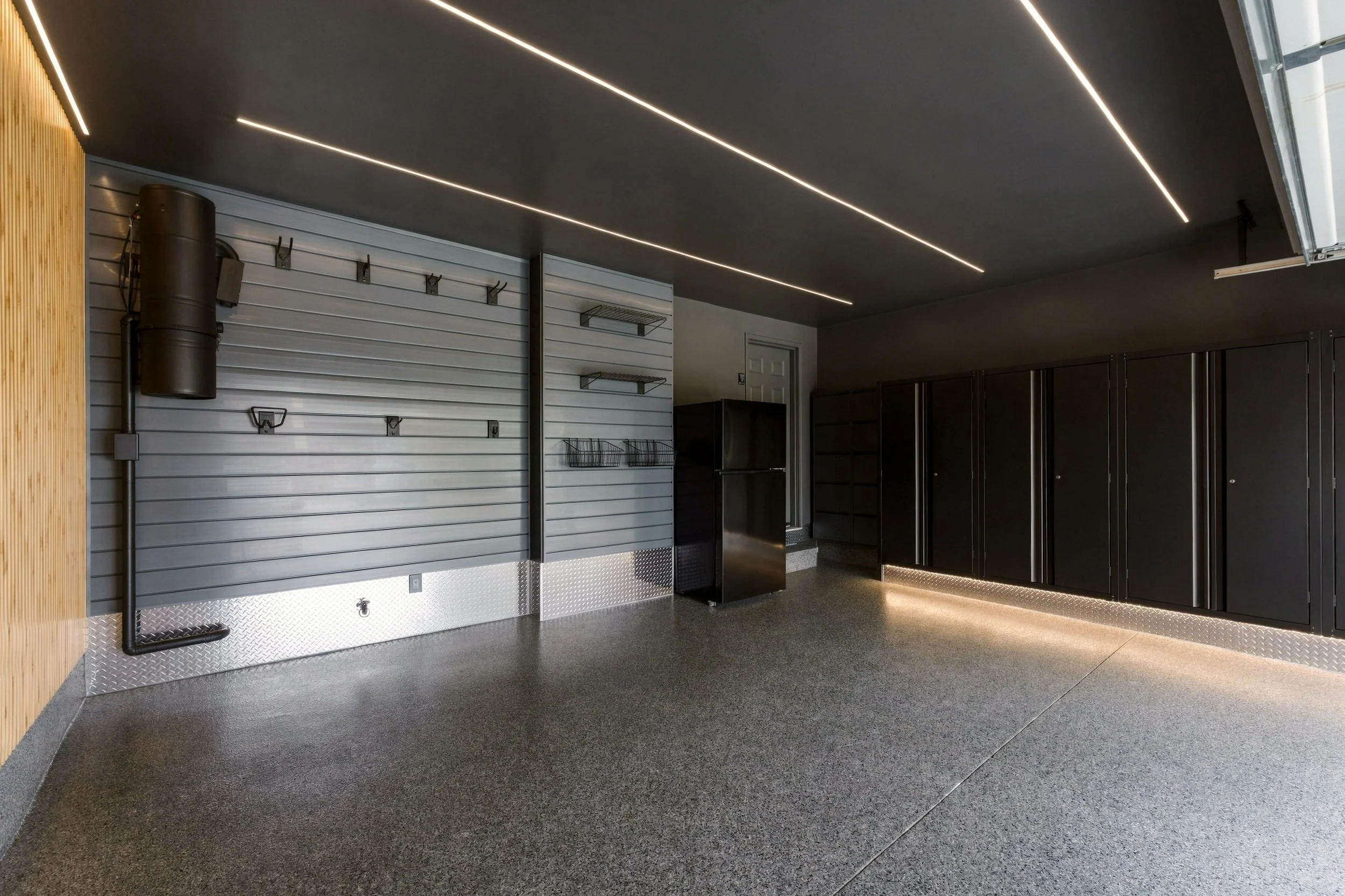 Modern garage interior with black ceiling, LED strip lighting, black cabinets, and a white slatwall with hooks and shelves.