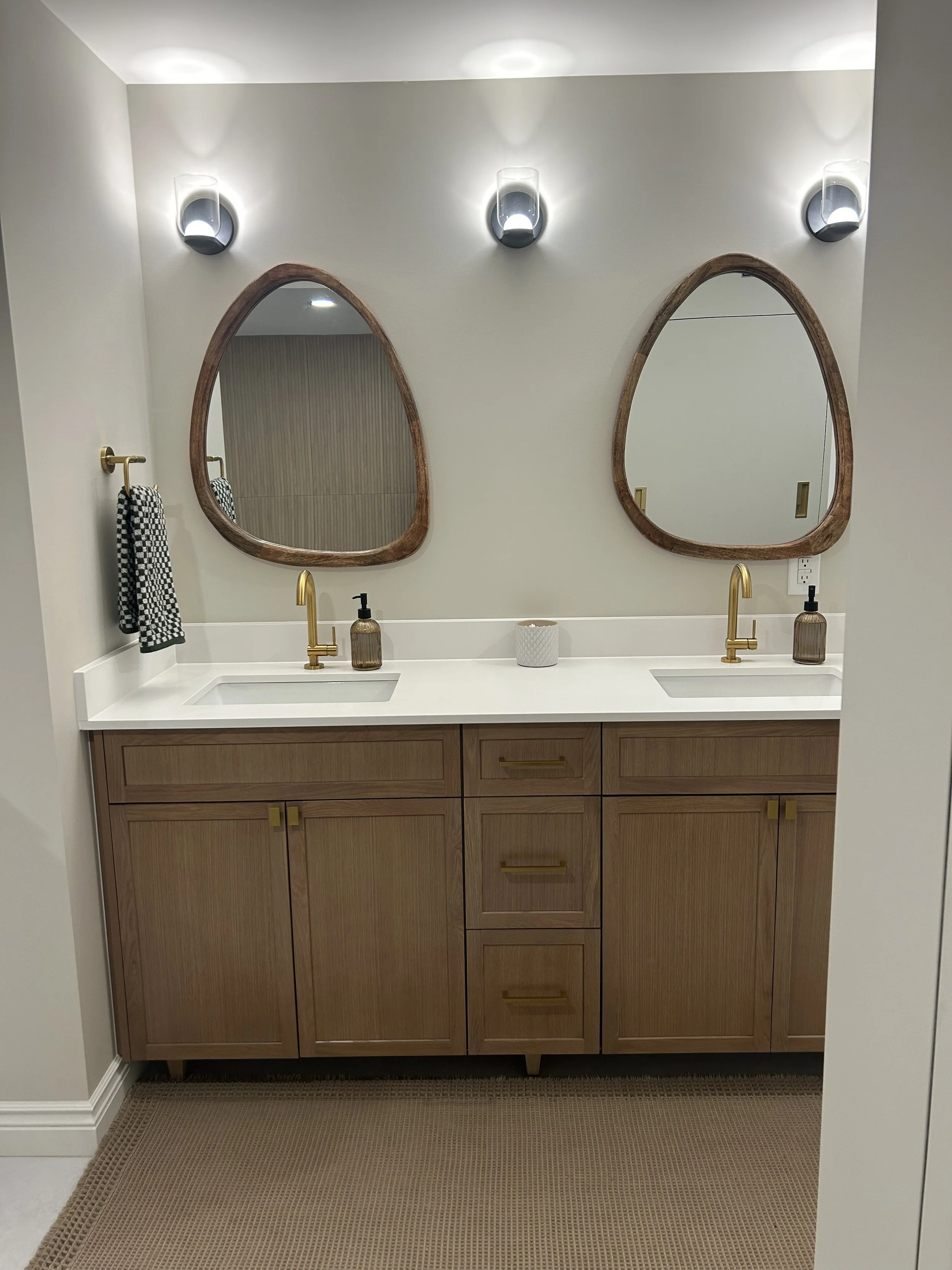 Bathroom vanity with two oval mirrors, two gold faucets, a black soap dispenser, and a white decorative container, set against a plain light-colored wall with three wall sconces.