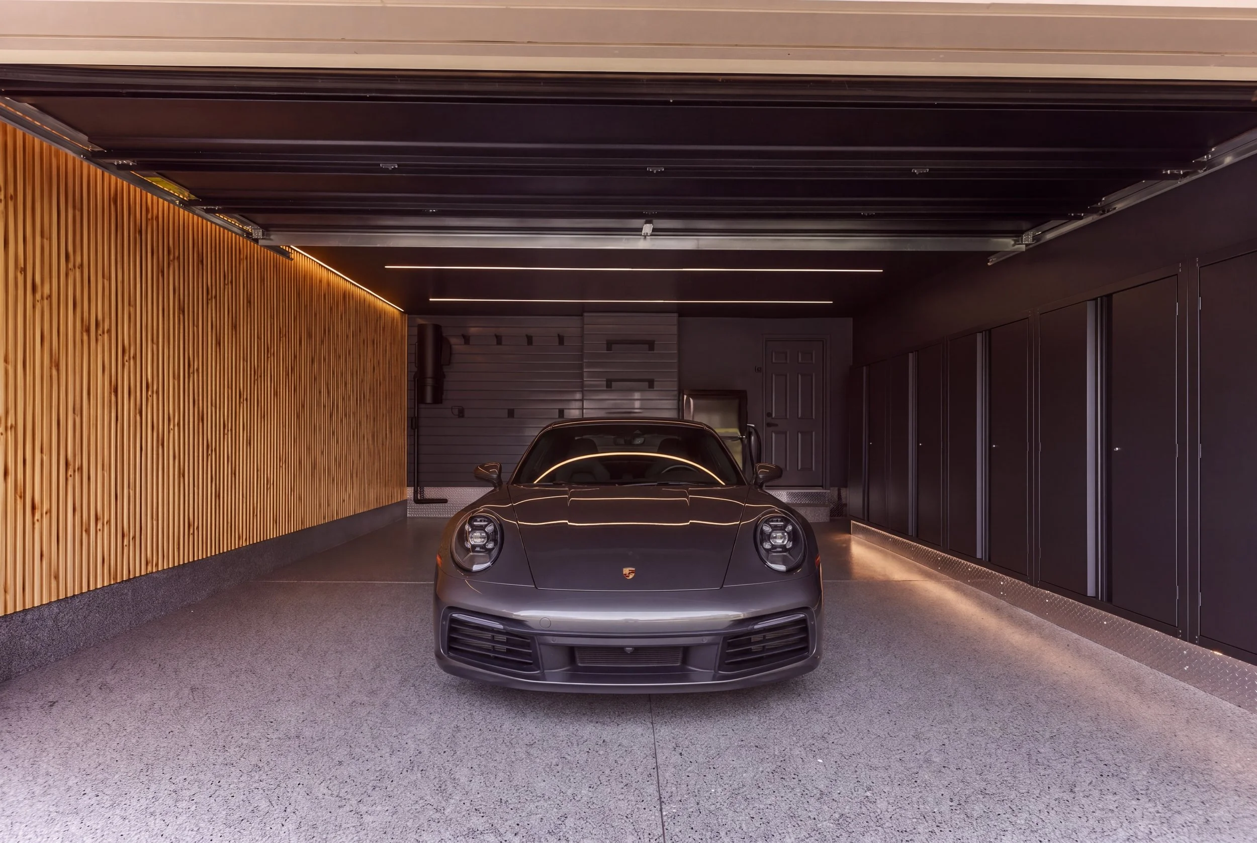 A sleek black sports car parked inside a modern garage featuring a wooden accent wall, black ceiling with integrated lighting, and black cabinetry along the right side.