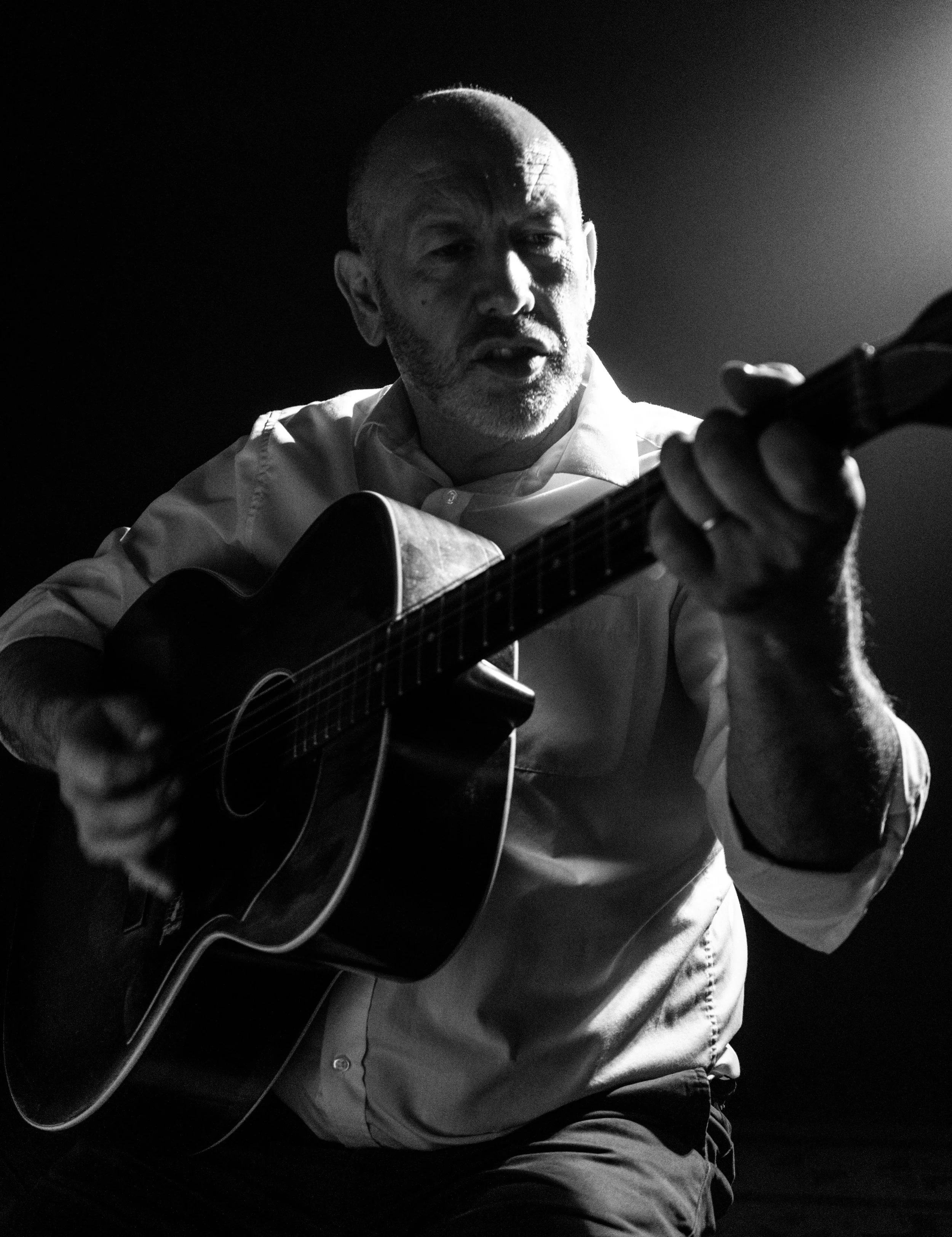 A black-and-white photo of an elderly man with a shaved head, playing an acoustic guitar. He is wearing a light-colored shirt, and his facial expression appears focused. The background is dark, emphasizing the man and his guitar.