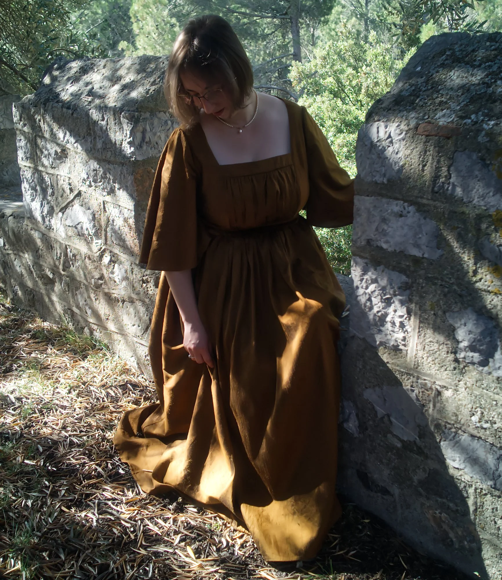 Une femme portant une robe marron et un collier en perle se tient entre deux murs en pierre dans une forêt ensoleillée.