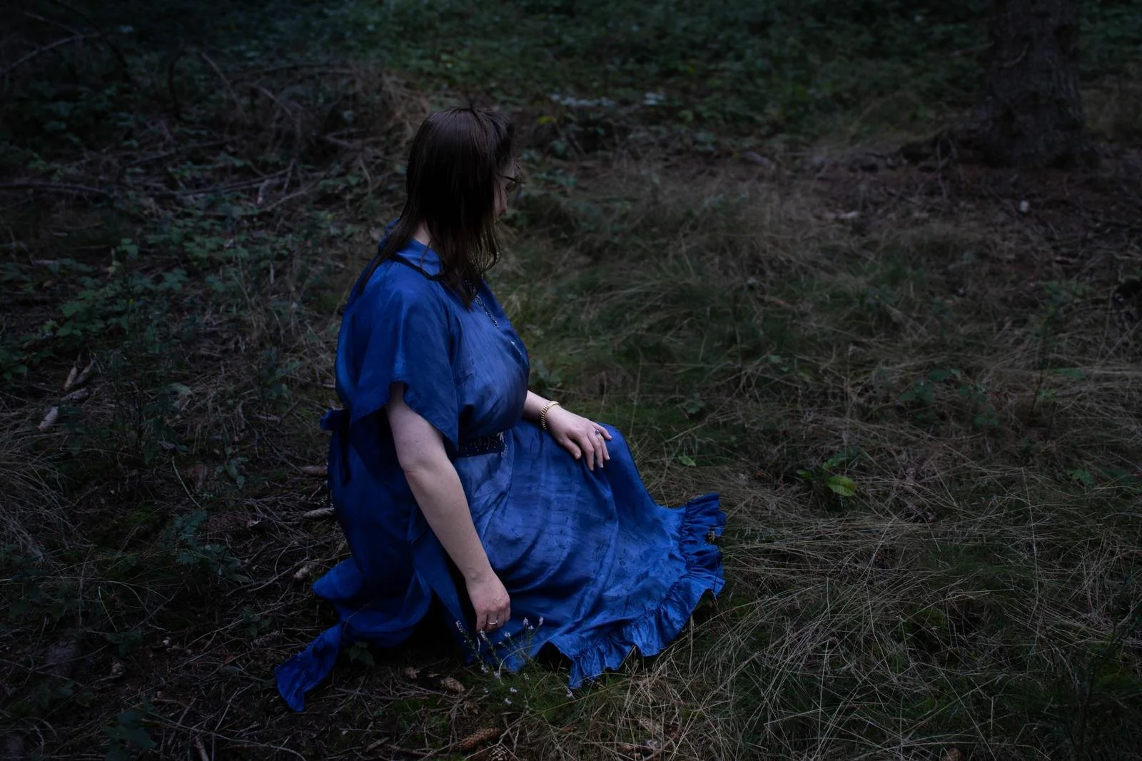 Femme assise dans une forêt, portant une robe bleue, regardant vers le sol.