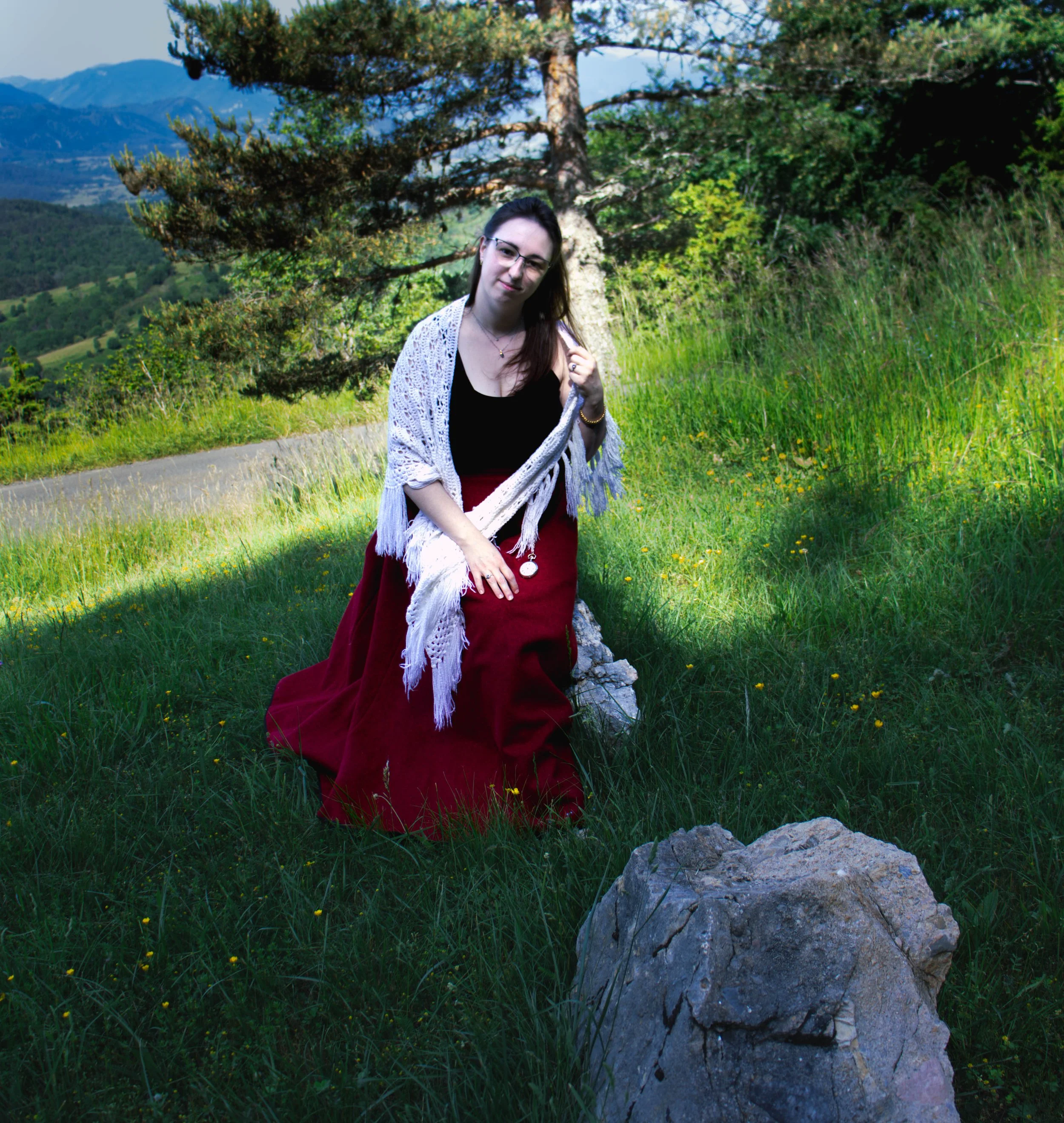 Jeune femme avec lunettes et foulard, assise près d'une grande roche dans un paysage naturel de montagne et forêt, en tenue mêlant élégance et style décontracté.