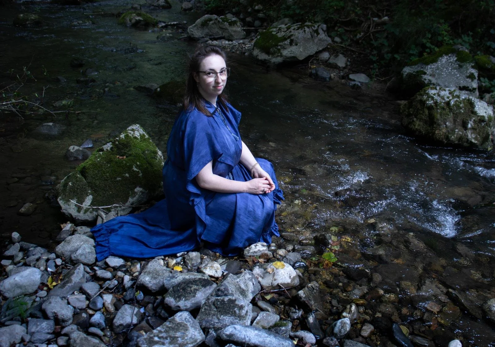 Une jeune femme vêtue d'une robe bleue, assise sur des rochers au bord d'une rivière dans une forêt.