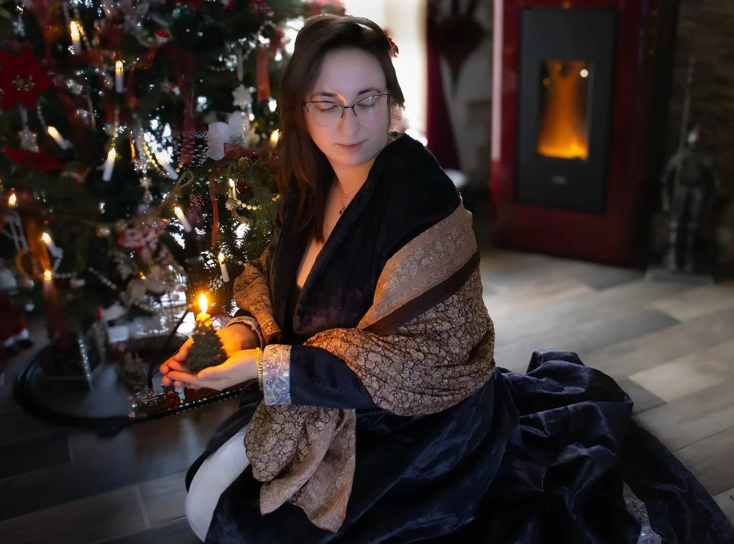 Une femme assise près d'un sapin de Noël décoré avec des guirlandes et des ornements, tenant une petite lanterne avec une bougie allumée, dans une pièce chaleureuse avec un poêle à bois en arrière-plan.