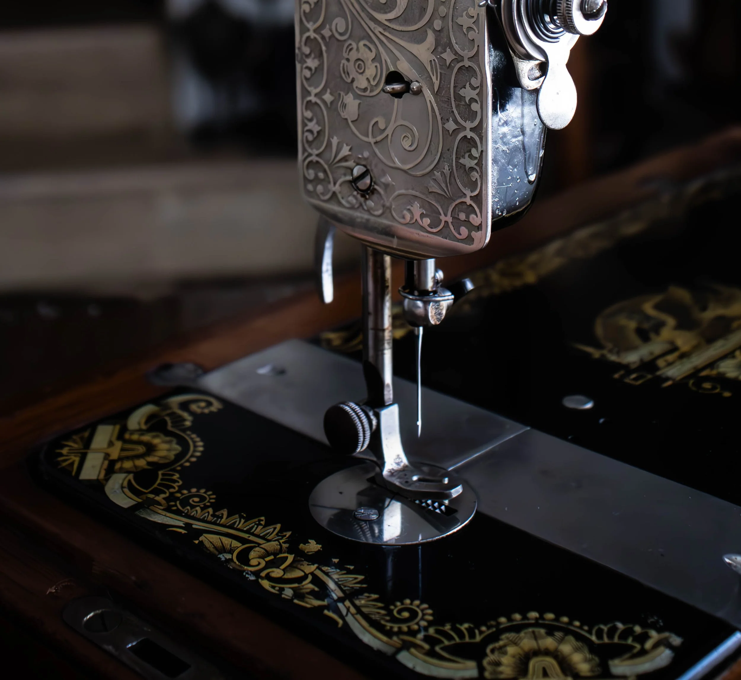 Détail d'une machine à coudre ancienne avec un décor en urée argentée, sur un fond noir avec des motifs dorés en bas.