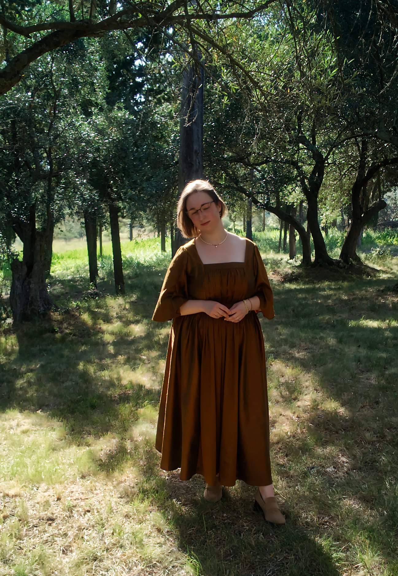 Une femme debout dans une forêt, portant une longue robe marron avec des manches courtes, des lunettes, un collier et une montre, sous un arbre.
