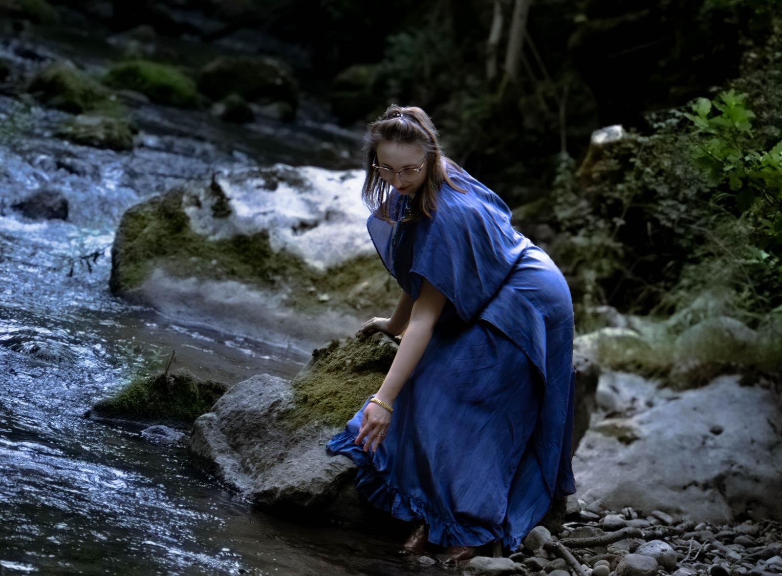 Une femme portant une robe bleue se tient sur des rochers dans une rivière entourée d'arbres.