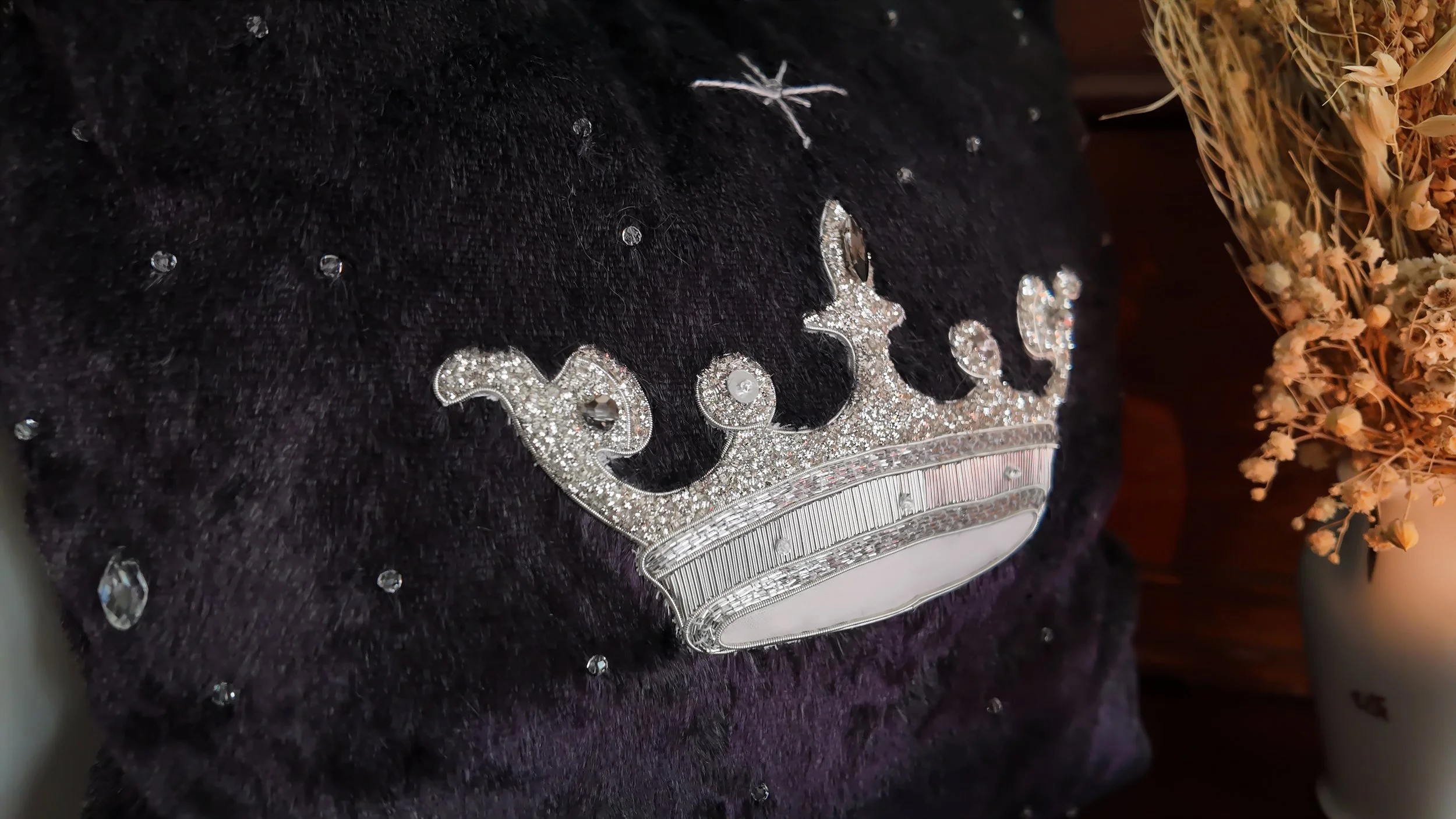 Une couronne en diamants posée sur un tissu noir en velours, avec des fleurs séchées en arrière-plan.