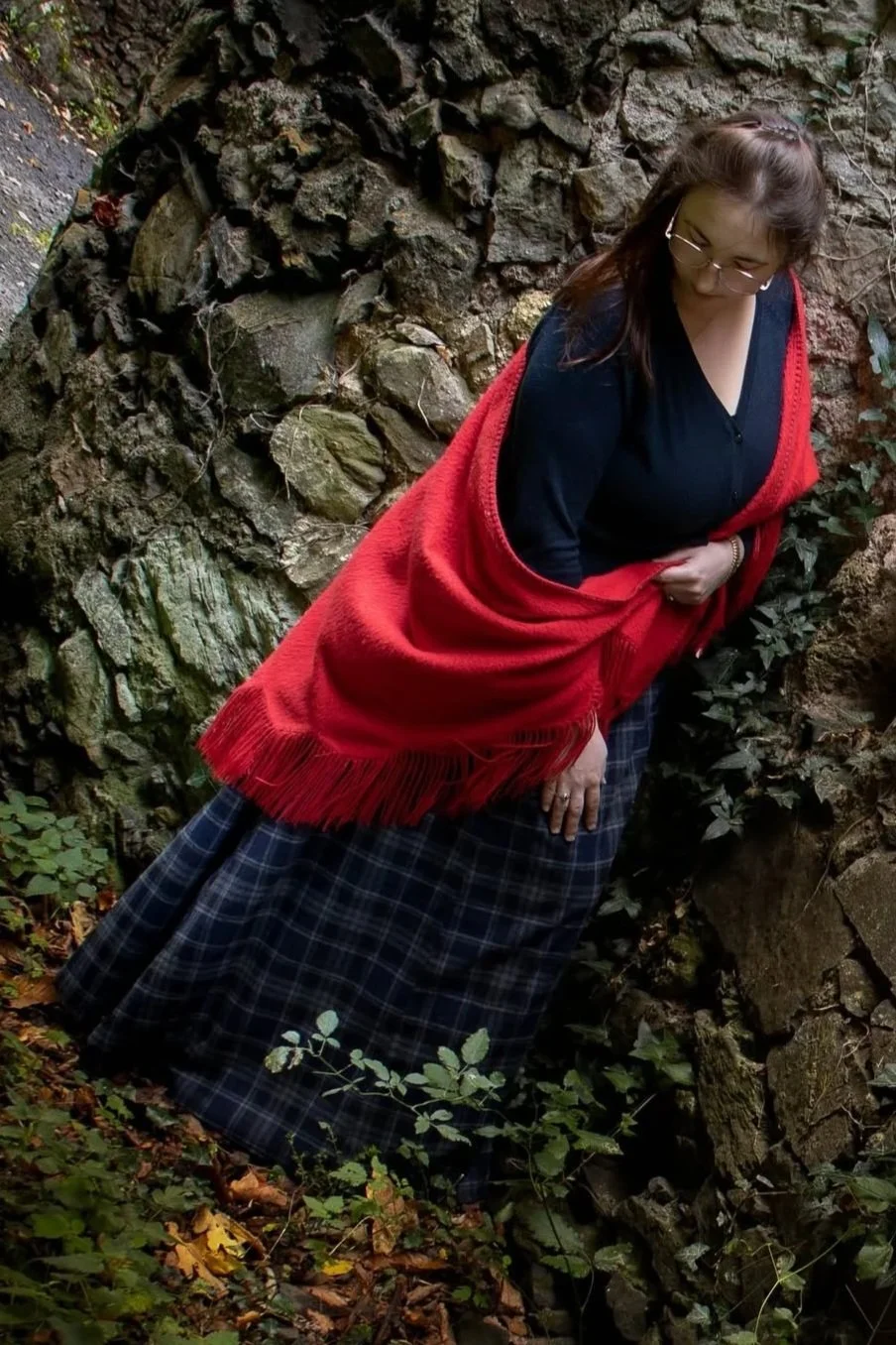 une femme portant une jupe à carreaux et une veste rouge, se tenant près d'une paroi rocheuse dans la nature.