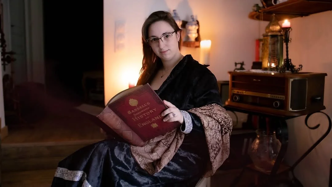 Une jeune femme avec des lunettes, assise dans une pièce chaleureuse, tient un livre rouge intitulé 'ASELL'S HISTORY OF ENGLAND'.