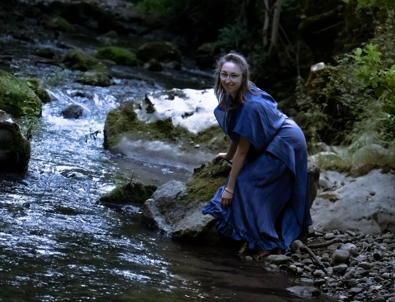 Une jeune femme portant une robe bleue posant près d'une rivière dans une forêt.