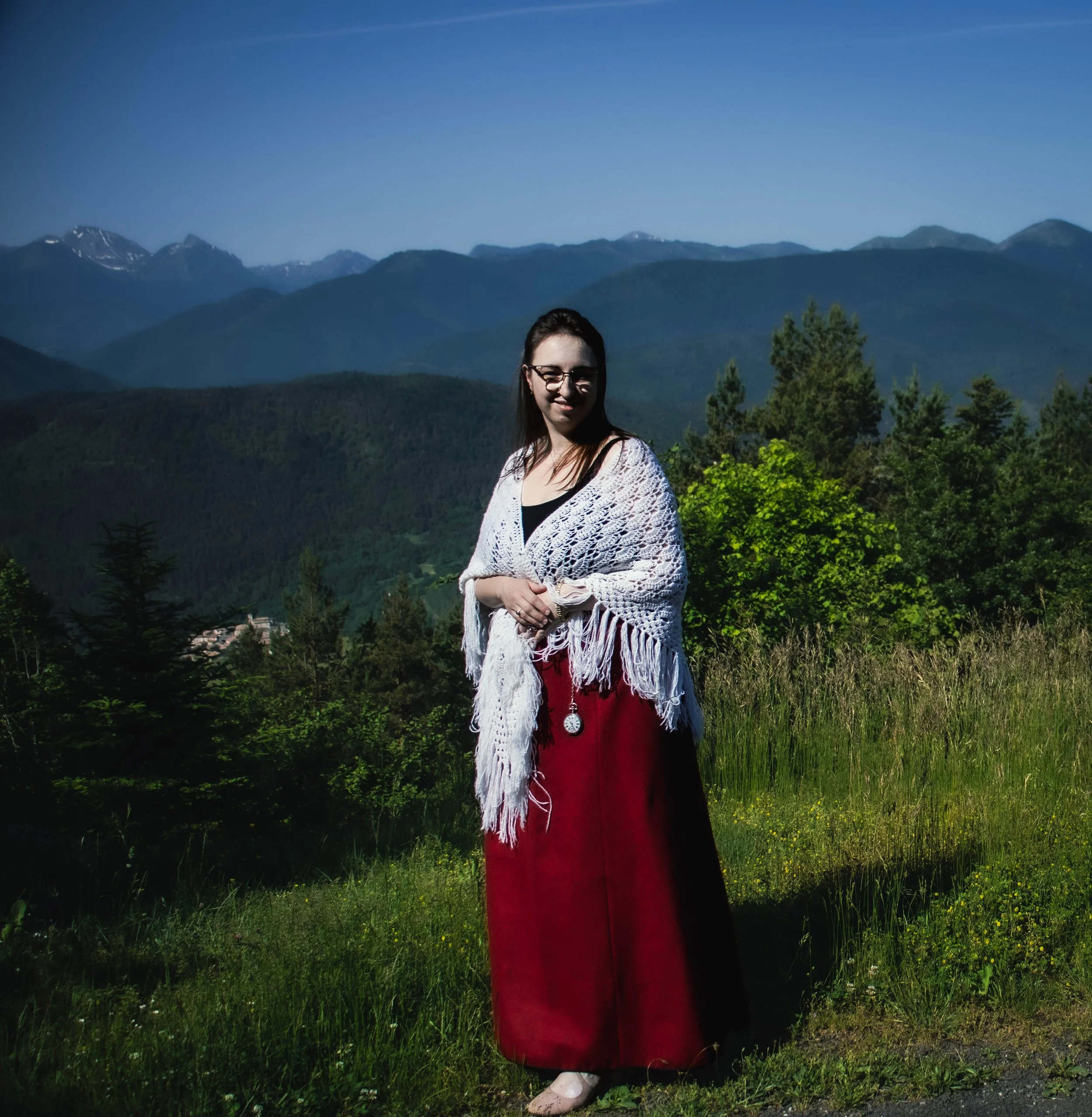 Une femme avec des lunettes, portant une robe rouge et une veste blanche en crochet, se tient dans un paysage de montagnes et de forêt.