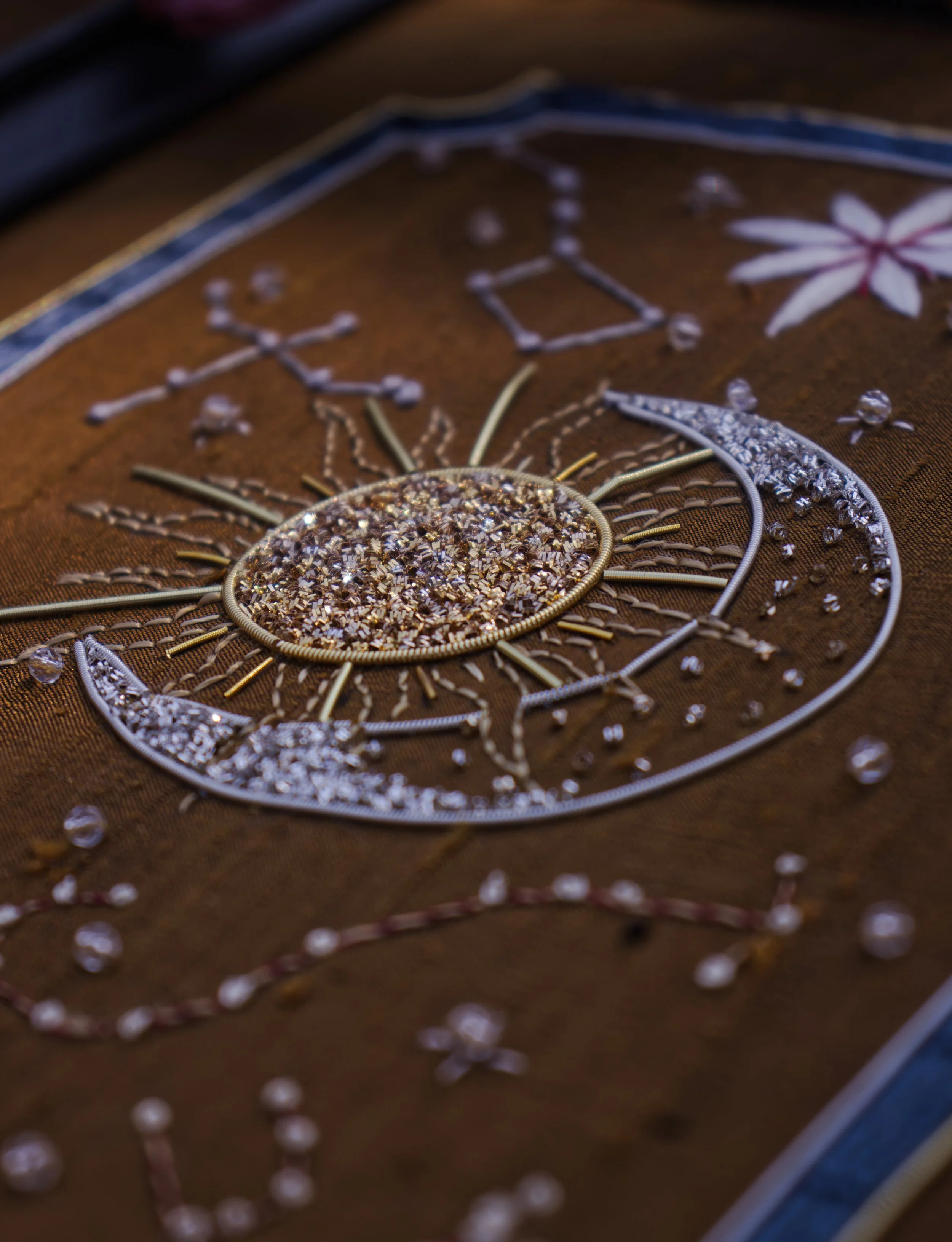 Une broderie en fil métallique, ressemblant à un soleil ou une fleur, sur un tissu marron avec des détails en pierres précieuses ou perles.
