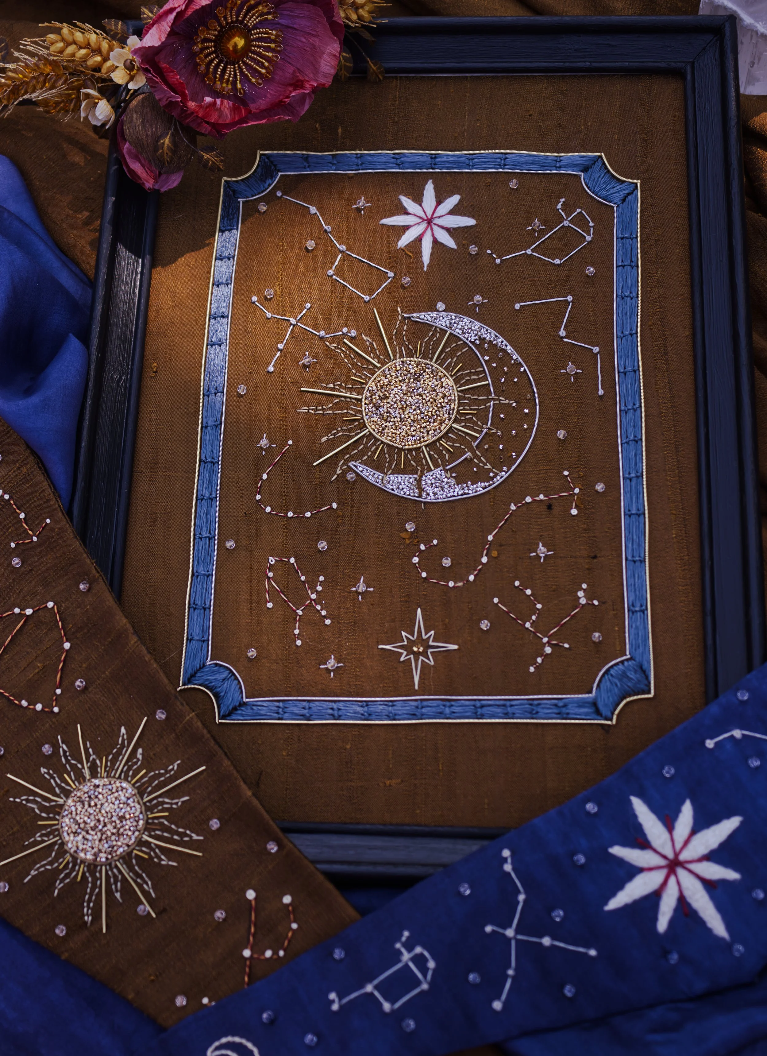 Une œuvre d'art avec des constellations, une lune, un soleil et une étoile, ornée de paillettes, de perles, et de rubans en forme de fleurs, dans un cadre en bois, entourée de tissus colorés.